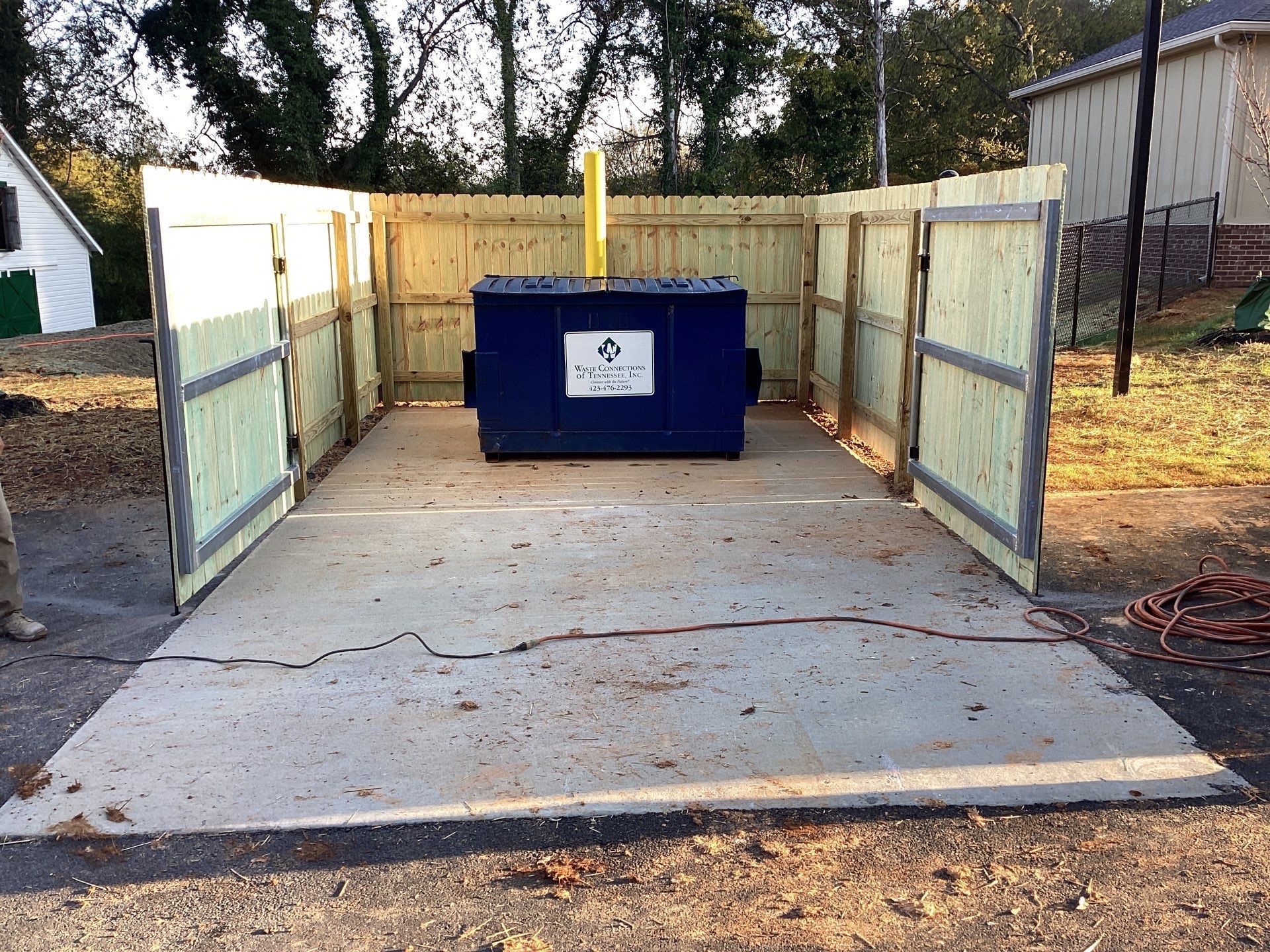 A blue dumpster enclosed by wooden fencing, on a concrete pad, outdoors.