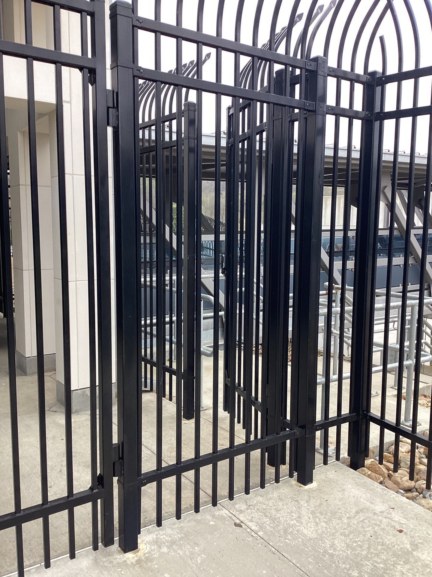Black metal gate with vertical bars and curved top; leads to a building with a concrete floor.