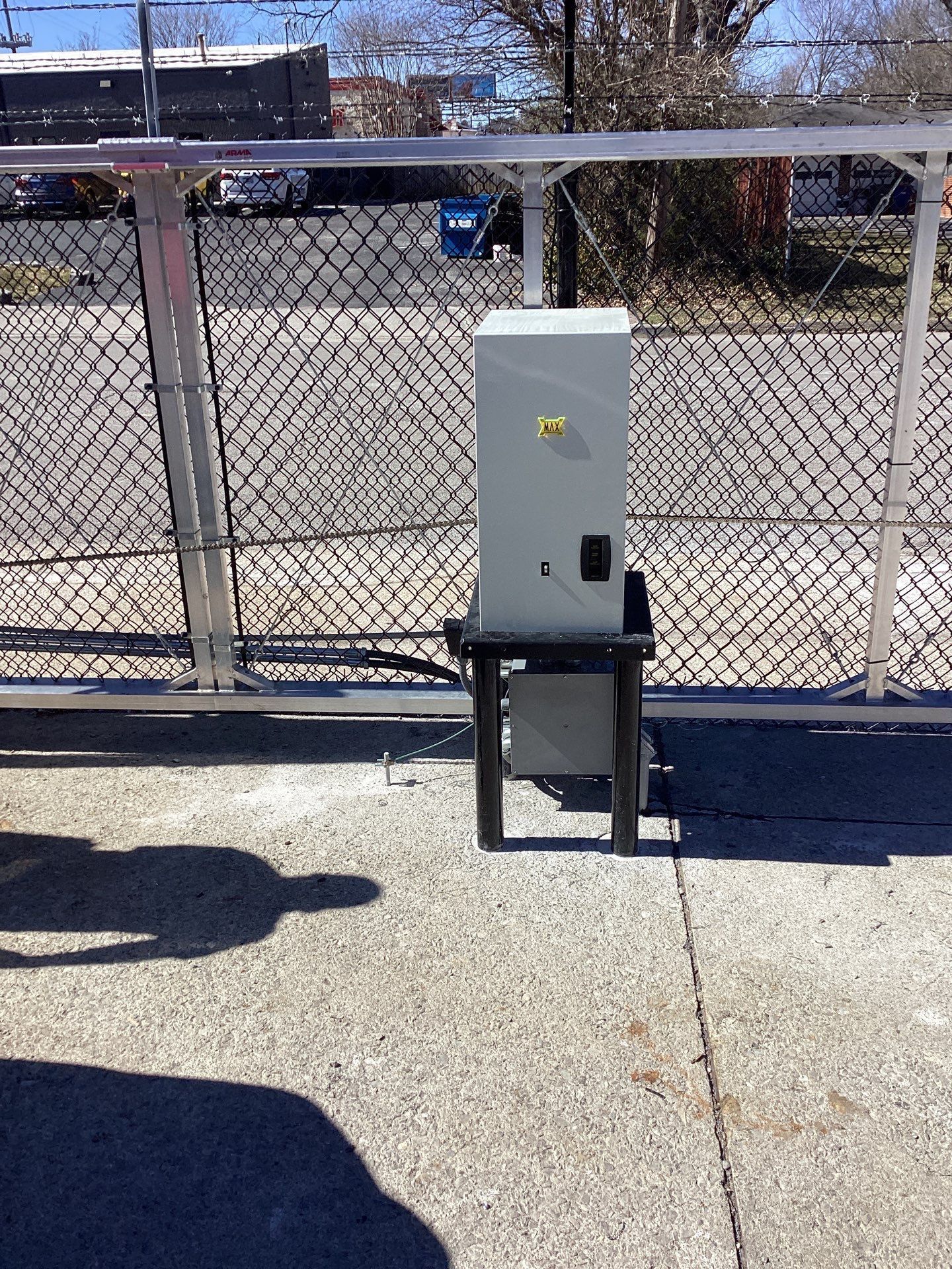 Gray control box on black stand next to a metal security gate. Sunny day, asphalt ground.