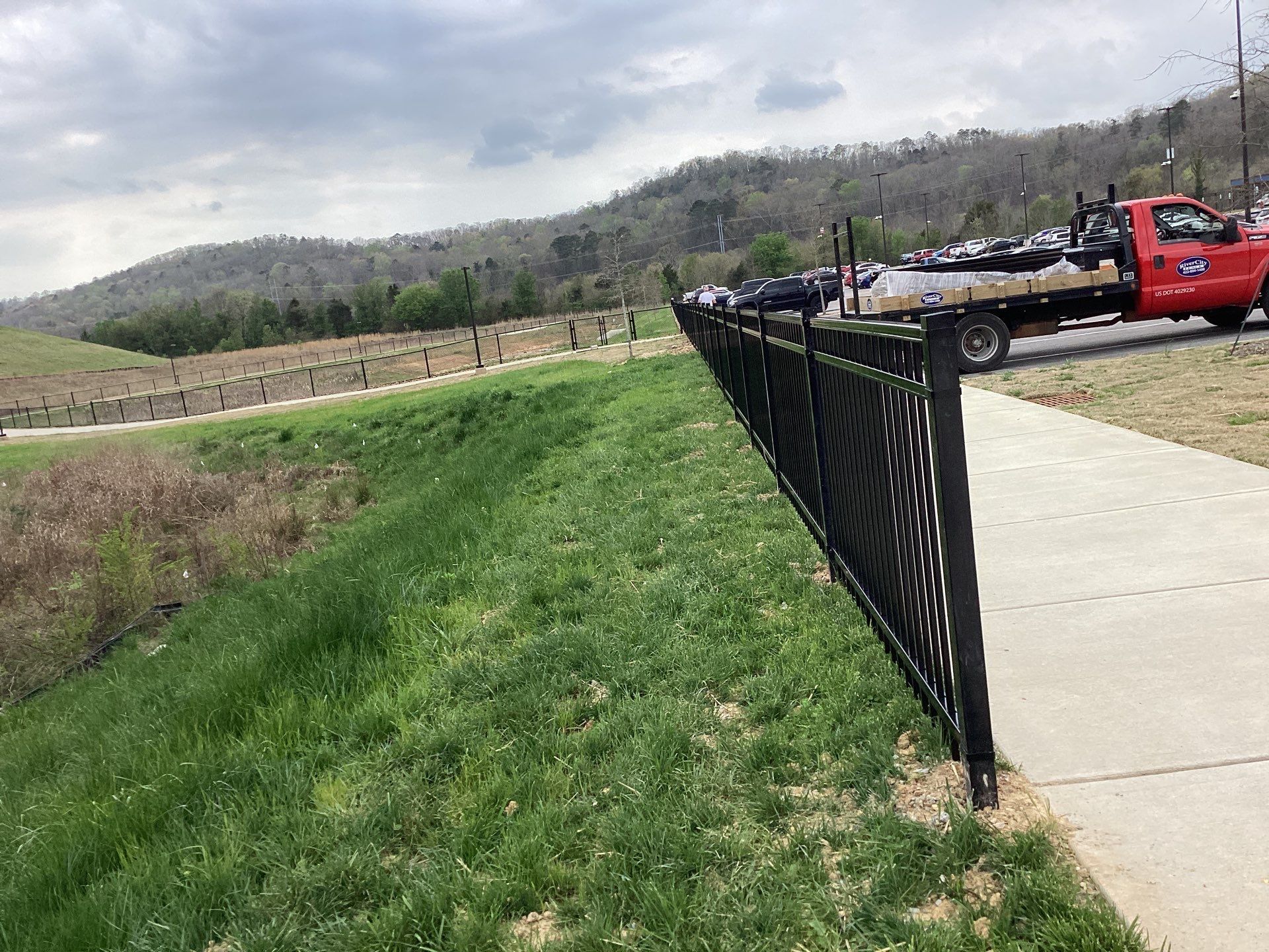 Black fence along a grassy slope and sidewalk, with a red truck and distant hills.