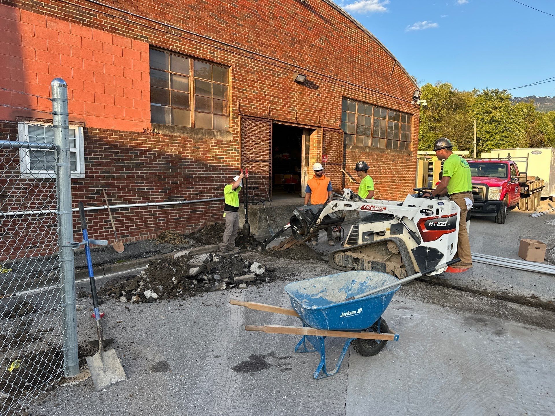 Construction workers outside a brick building, using equipment. A wheelbarrow holds material.