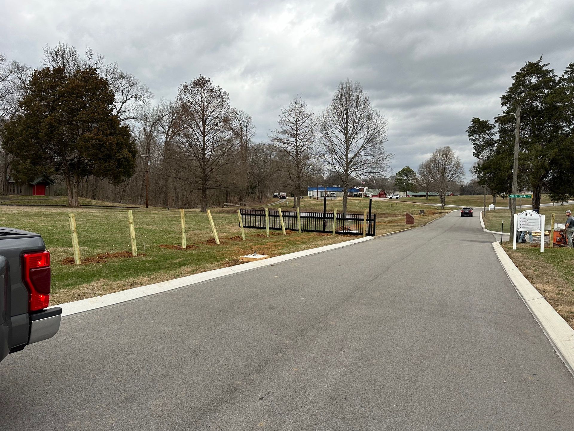 Paved road leading into a park on a cloudy day. Trees, fences, and parked vehicles visible.