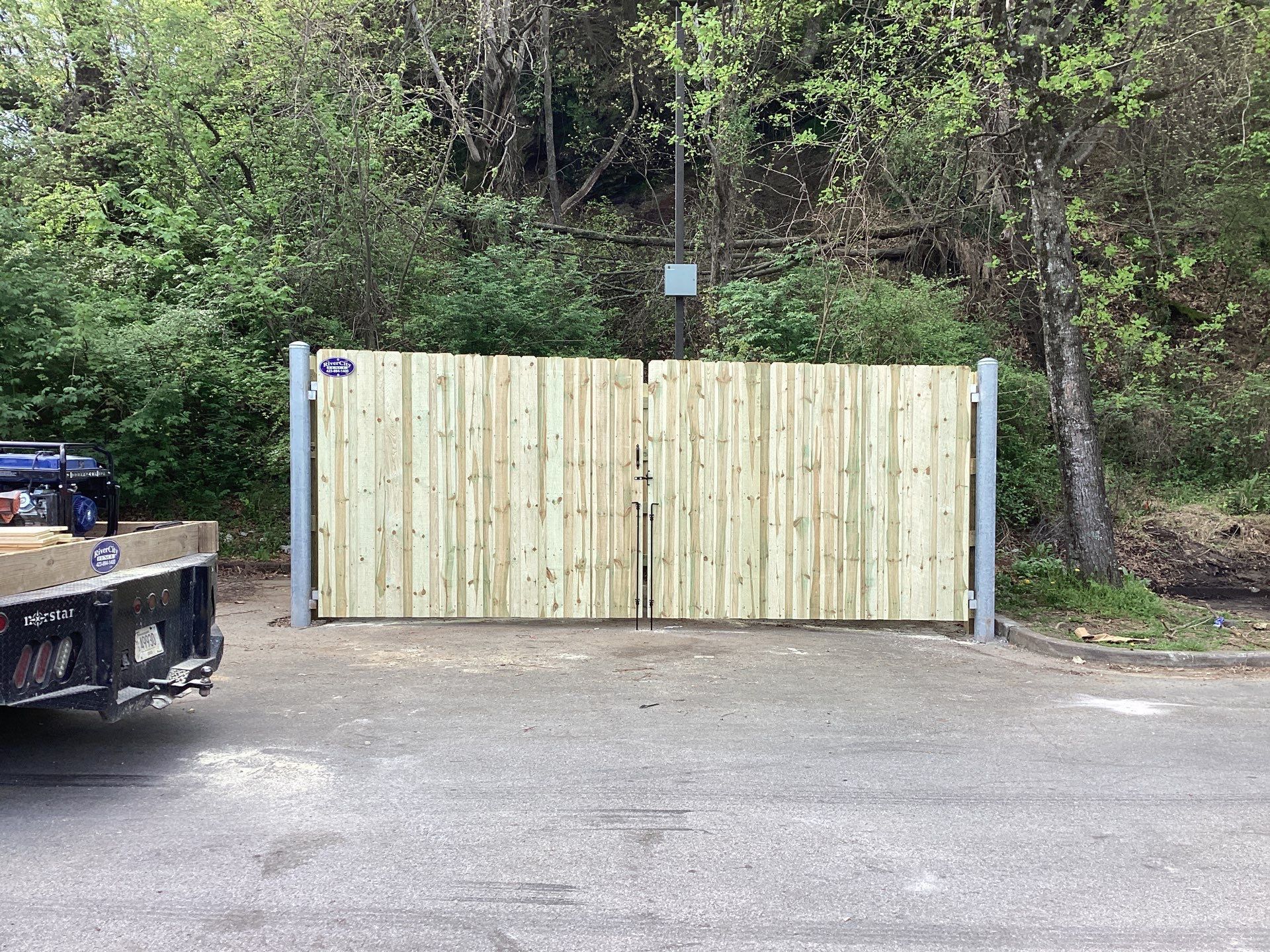 Wooden double gates on gray metal posts in front of a hillside covered in trees and bushes.