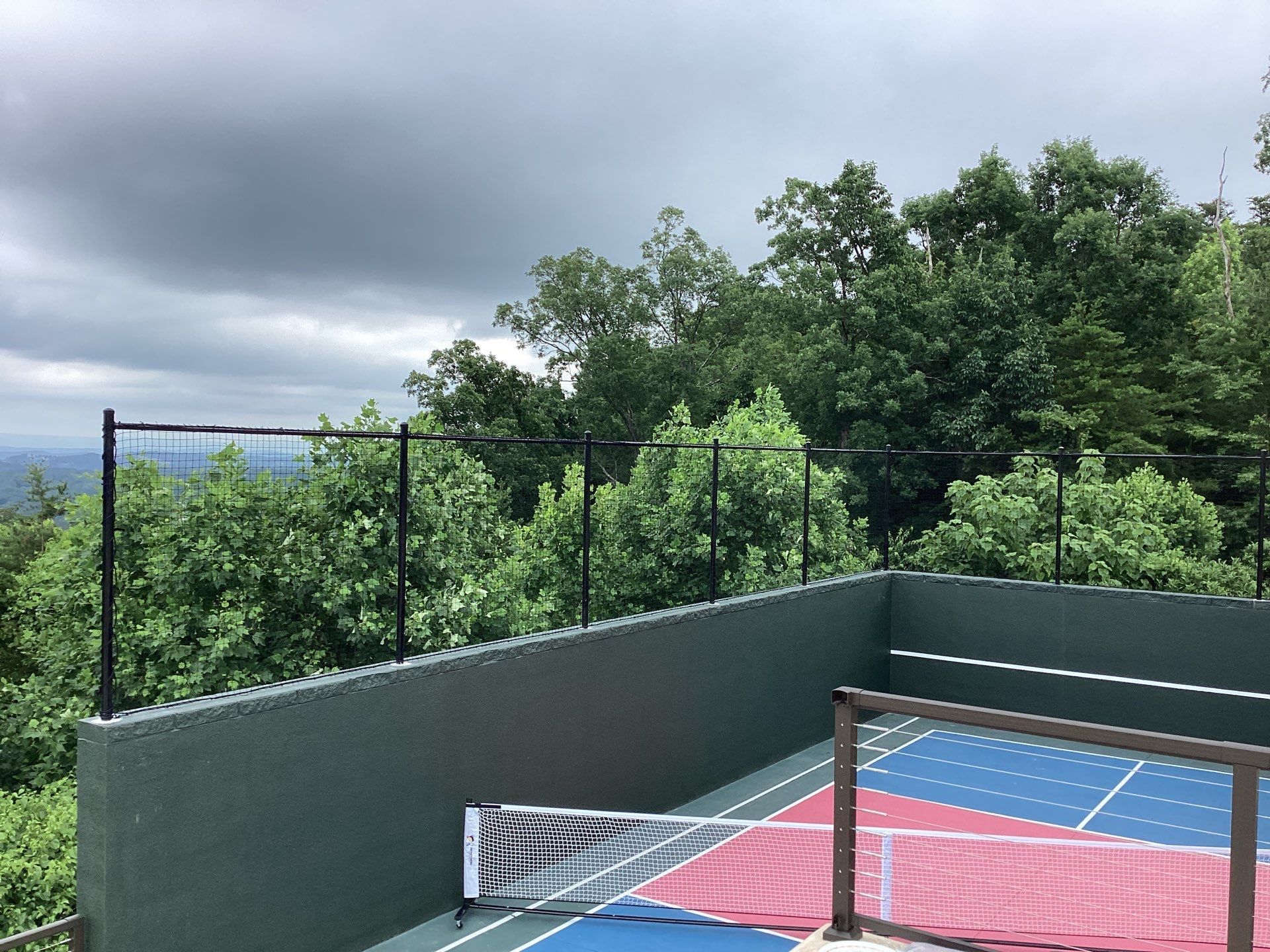 Pickleball court with net and colorful surface, overlooking trees and a cloudy sky.