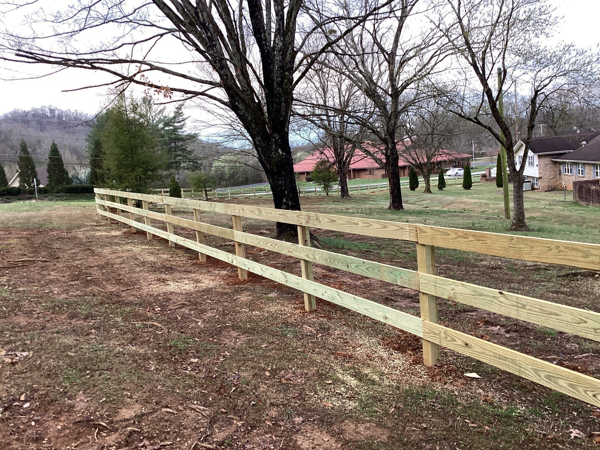 Wooden rail fence in a grassy field with trees and a house in the background.