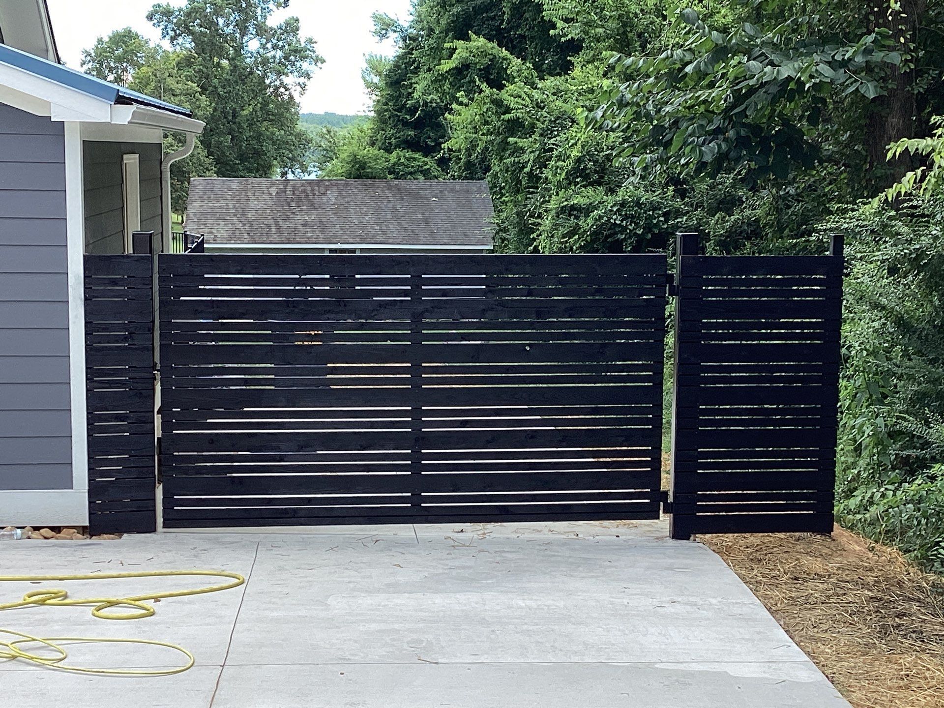 Black horizontal slat driveway gate and fence on a concrete driveway, with greenery in the background.