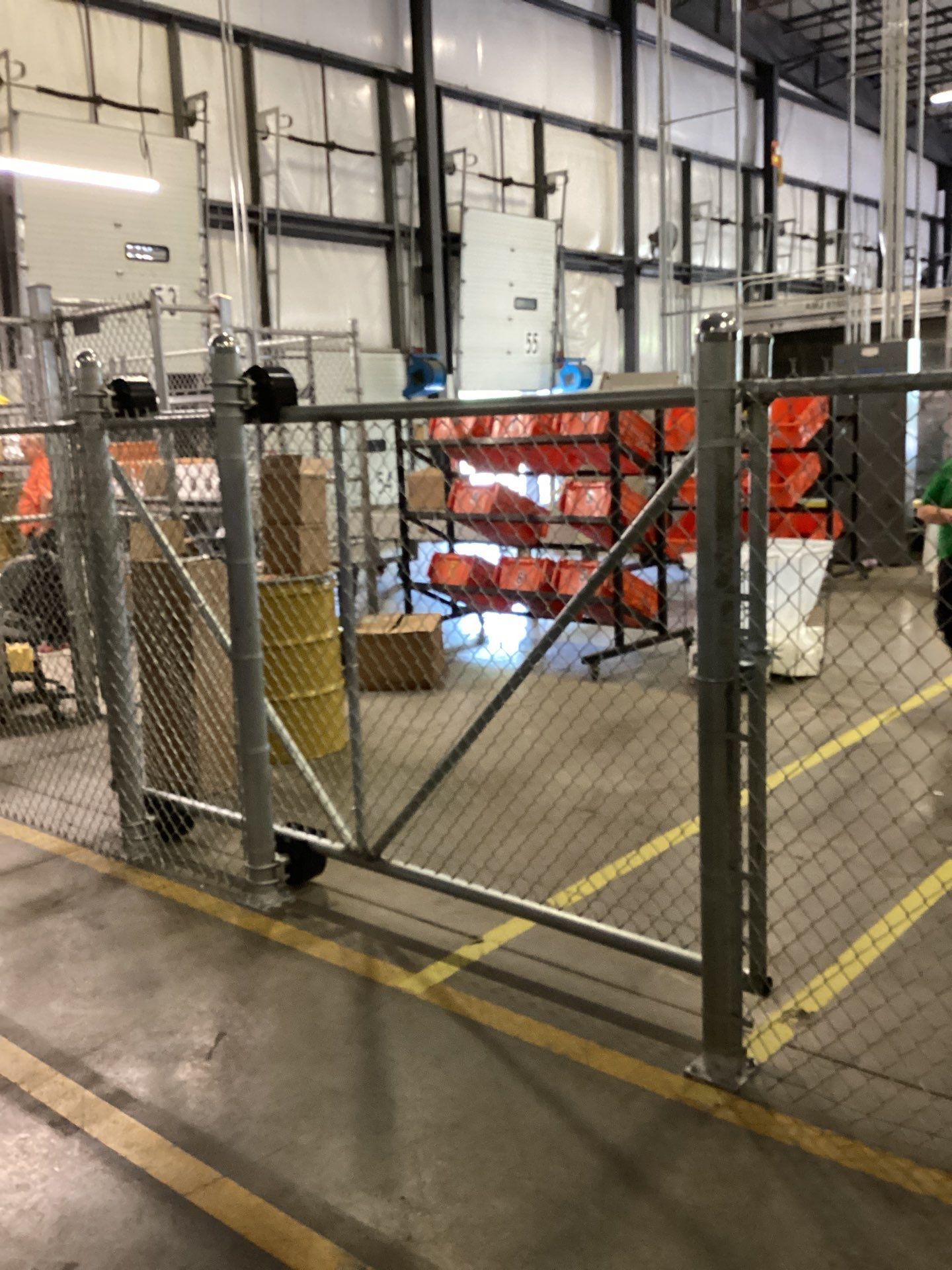Chain-link gate inside a warehouse, with boxes, barrels, and orange storage bins visible in the background.
