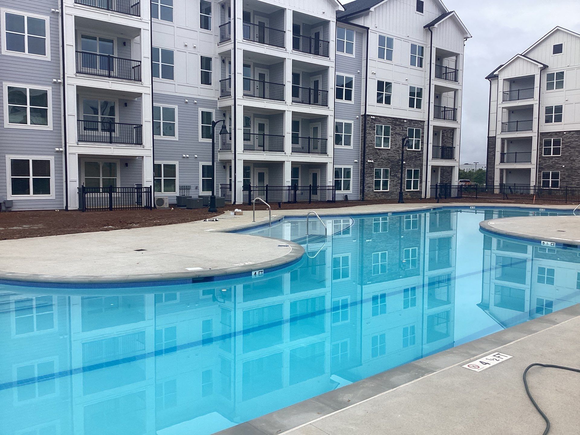 Swimming pool in front of multi-story apartment buildings with balconies. Buildings have gray siding and stone accents.