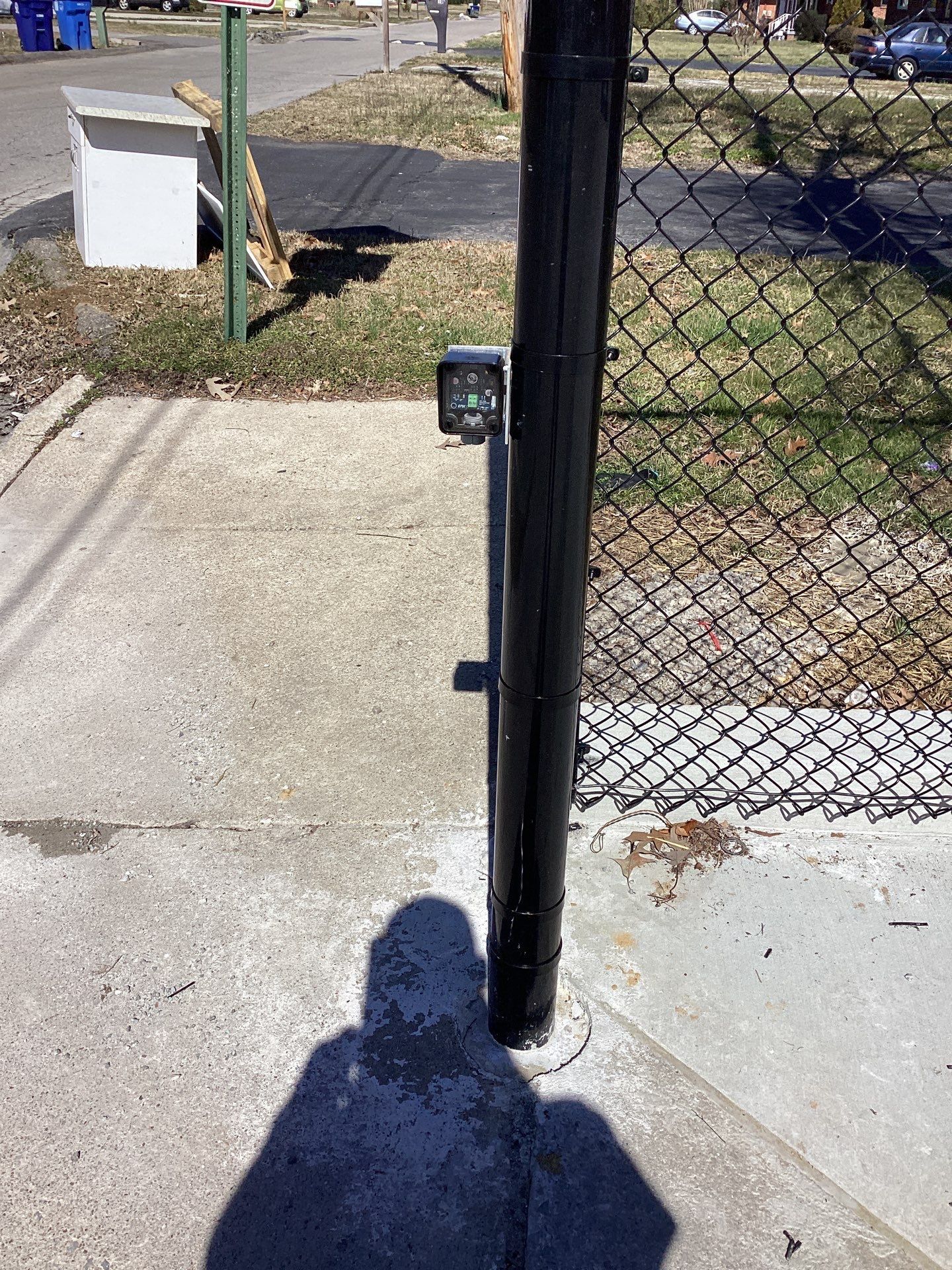Black metal gate post with access control panel, on a sidewalk, adjacent to a chain-link fence.
