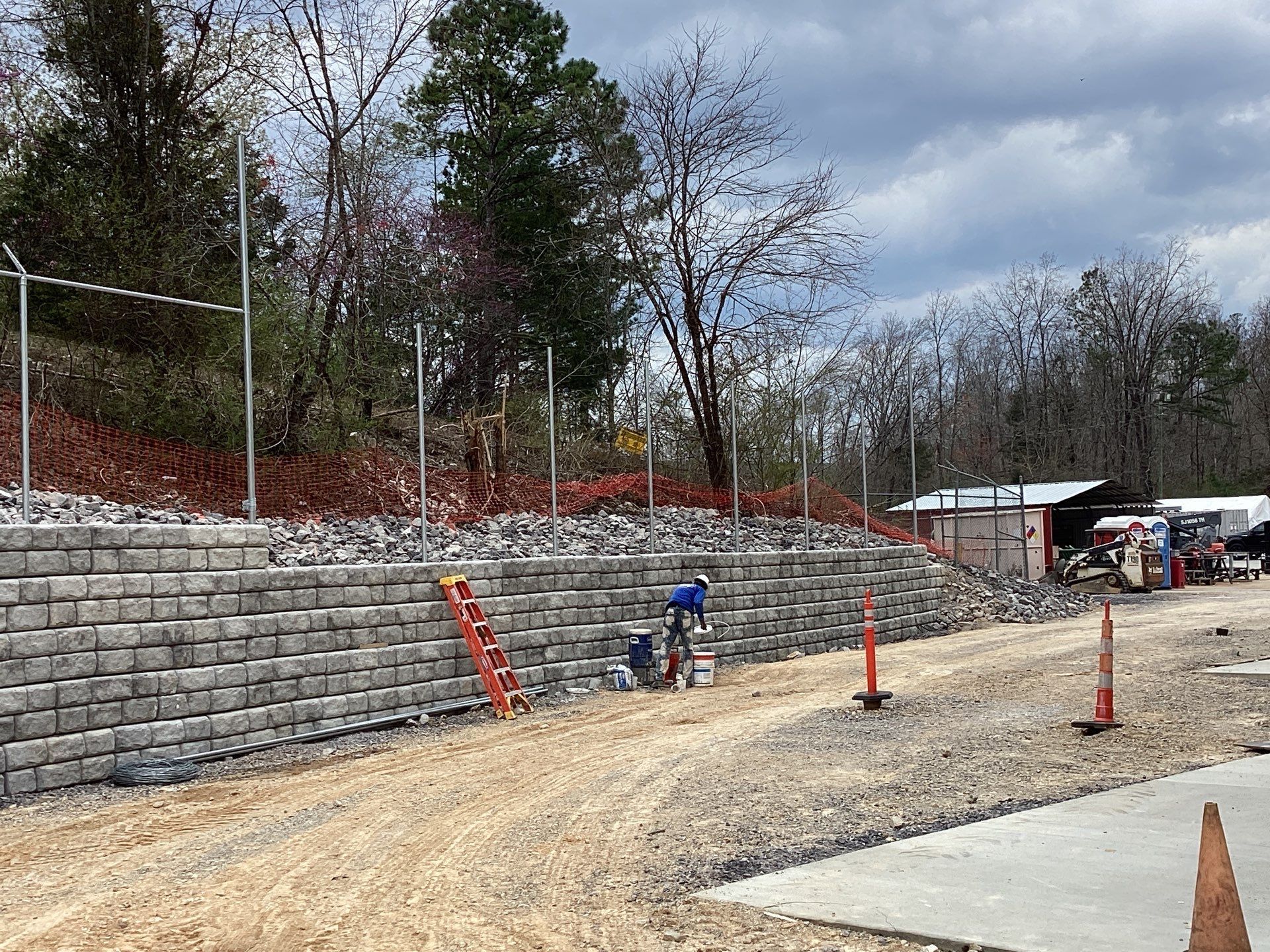 Construction site with retaining wall, dirt path, safety cones, and equipment under cloudy sky.
