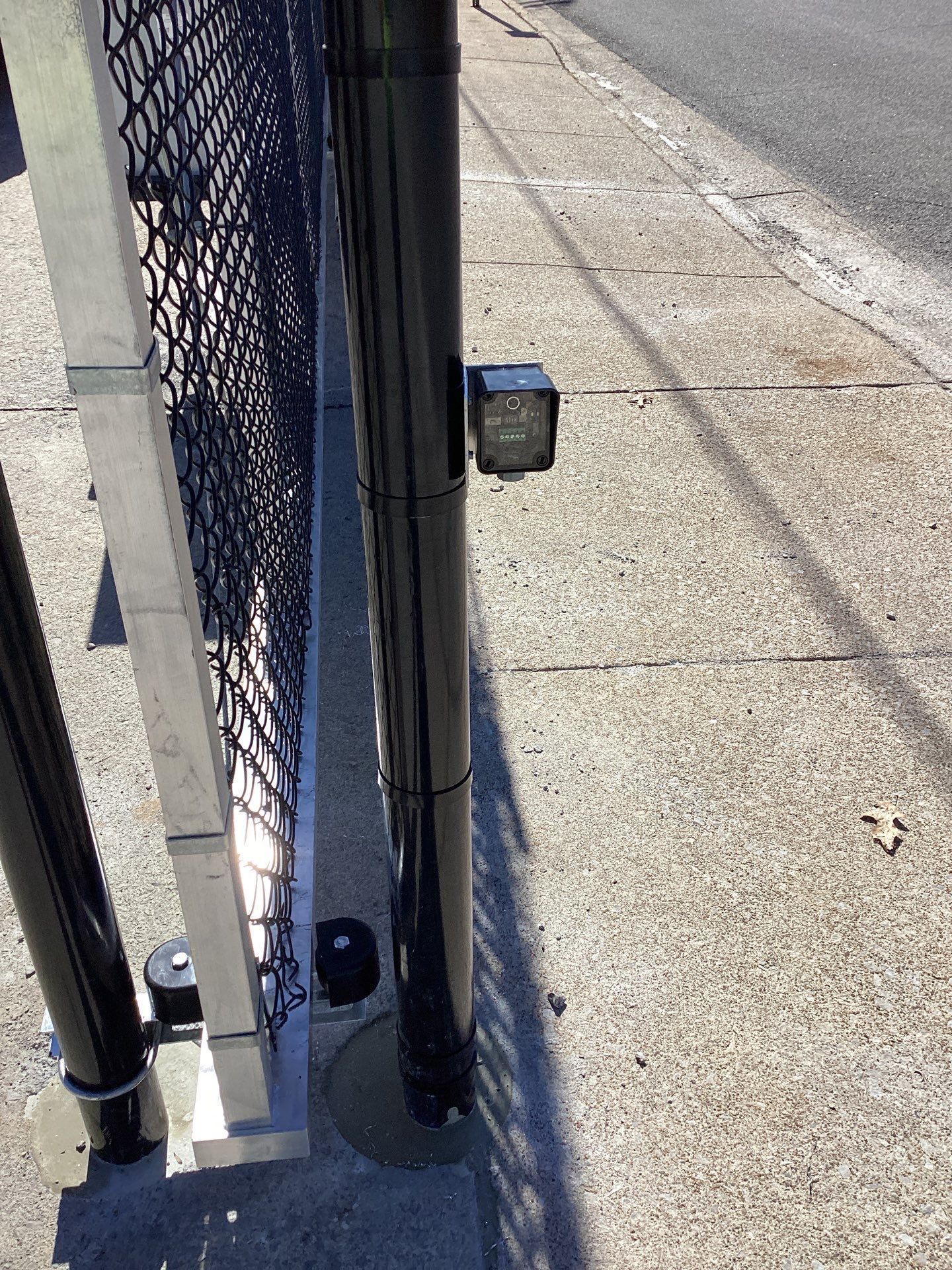 Black security camera mounted on a black pole next to a chain-link fence and concrete sidewalk.