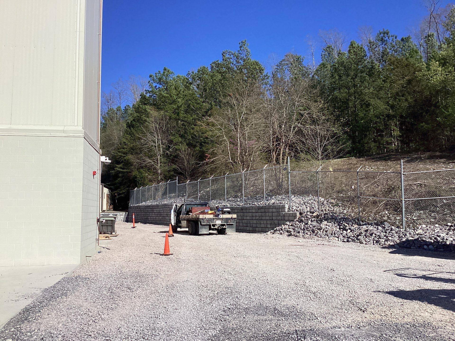 Gravel lot with building, fence, and trees. A truck is parked near the fence, with traffic cones present.