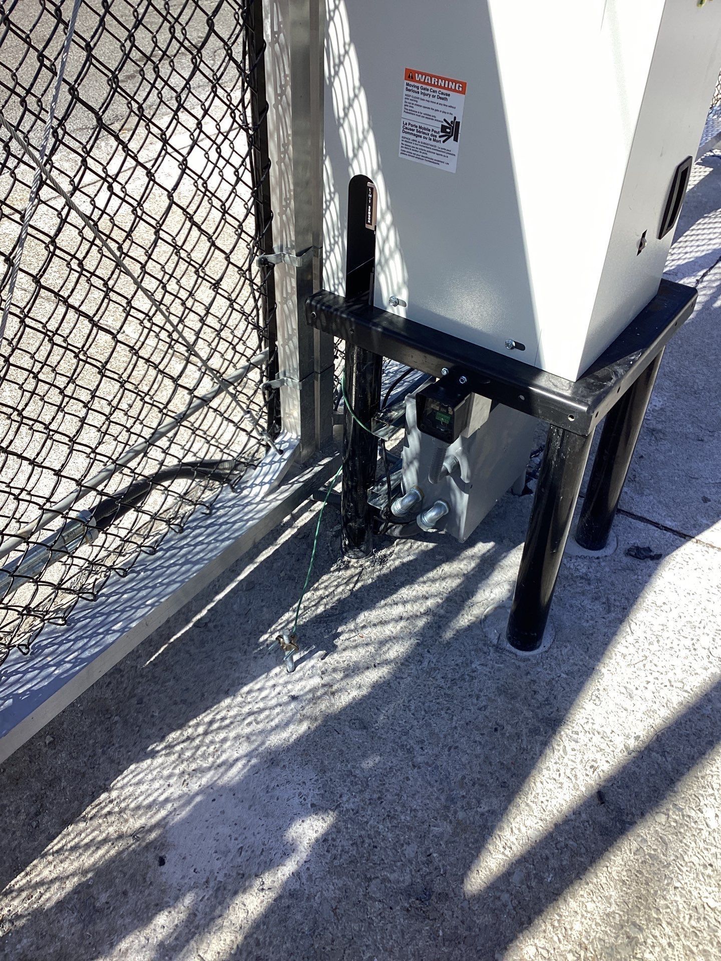 Metal electrical box mounted on a black metal stand next to a chain-link fence on concrete.