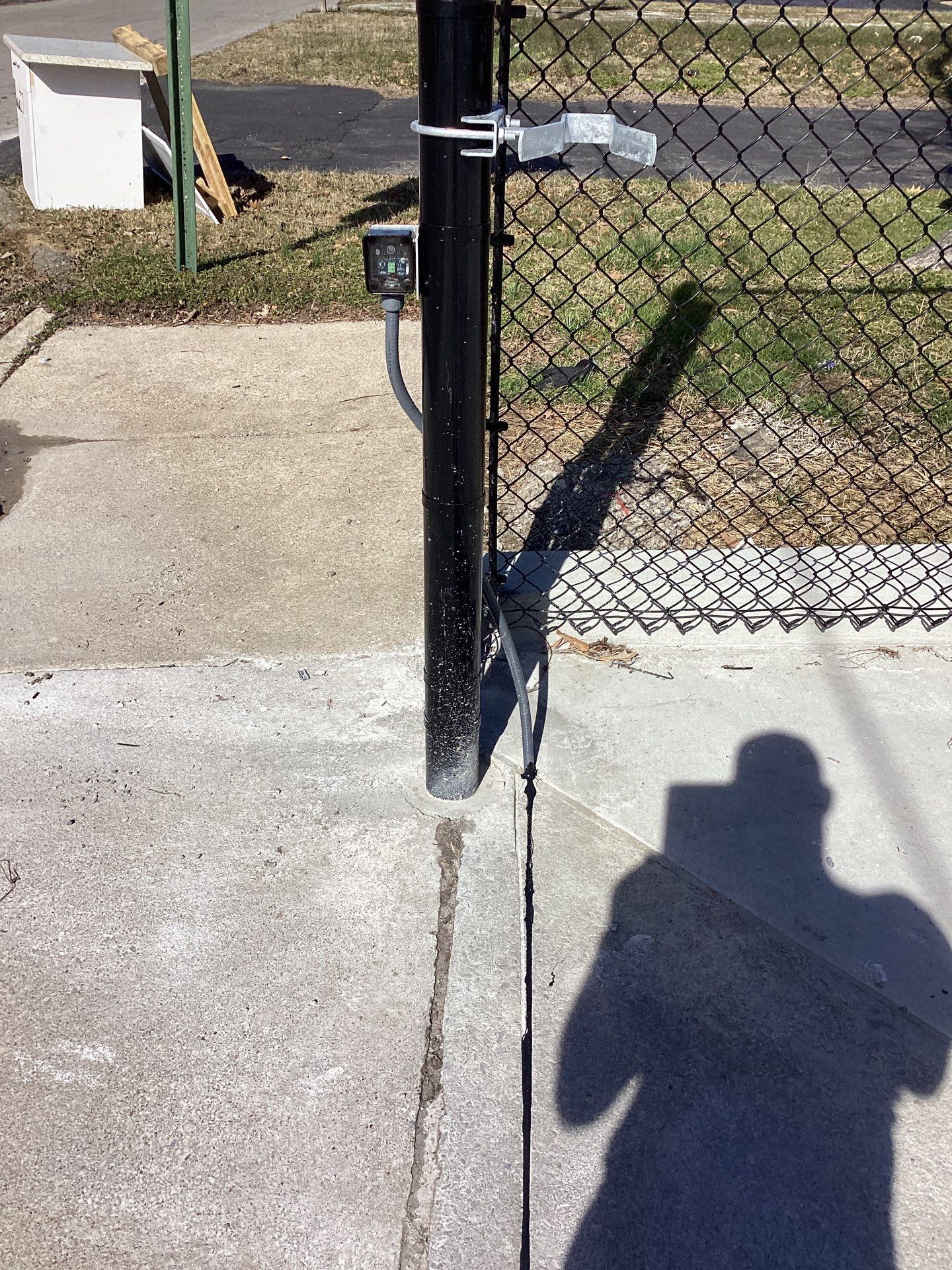 Black metal gate with a latch, sensor, and concrete walkway. Shadow of a person taking the picture.