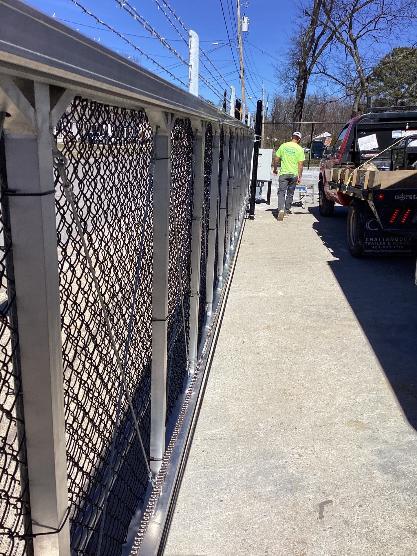 Silver metal chain-link fence with barbed wire on top; a person walks near a truck parked alongside it.
