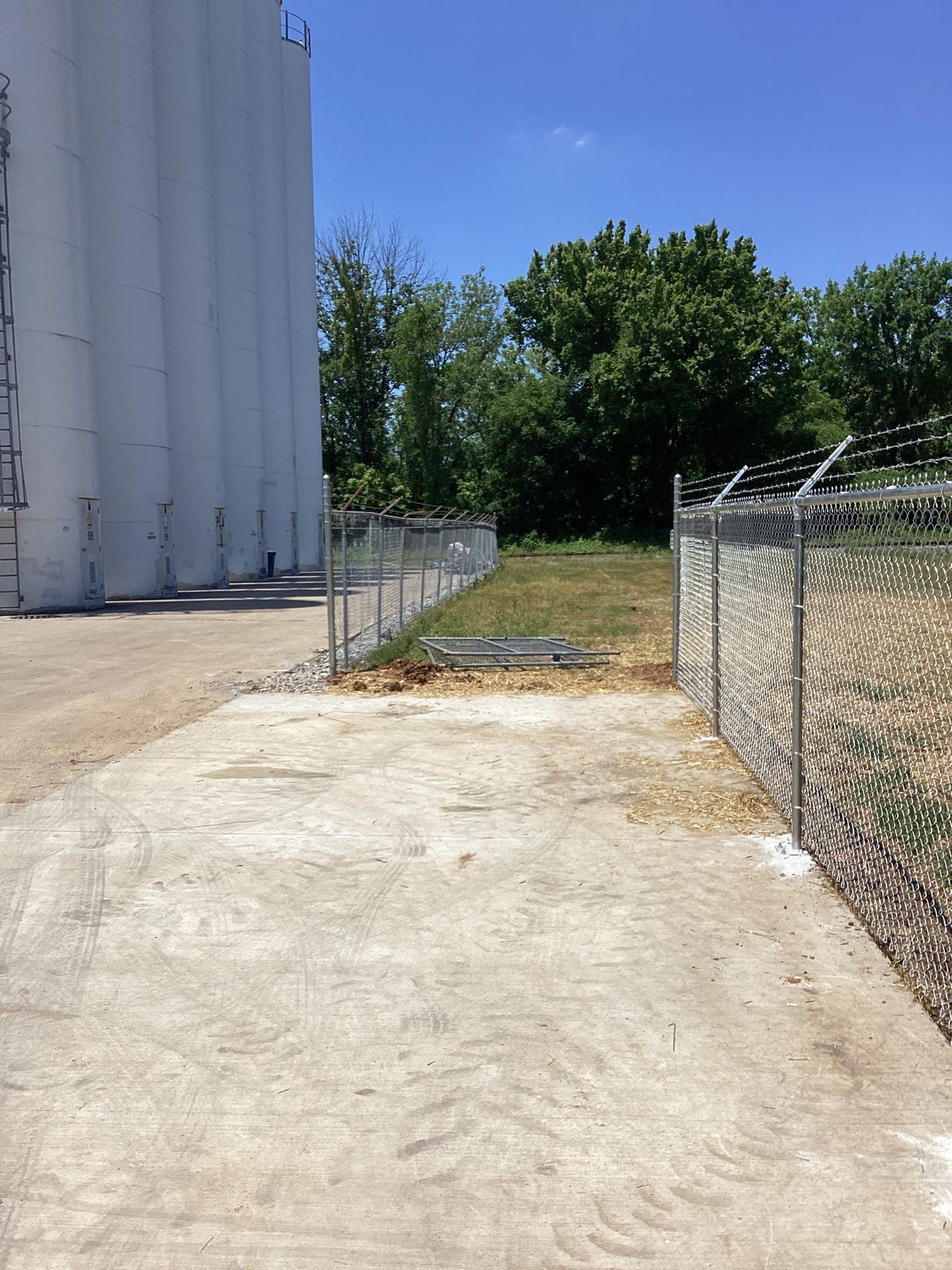 Chain-link fence with barbed wire surrounds a grassy area next to a large white industrial structure under a blue sky.