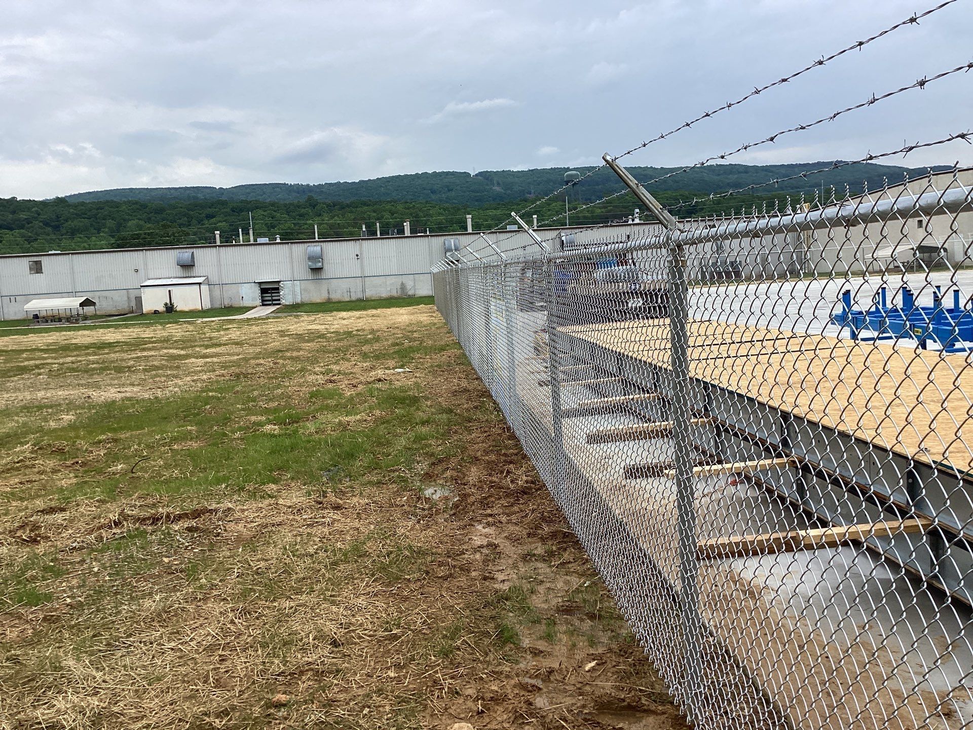 Chain-link fence with barbed wire. Industrial building and green hill in the background. Overcast sky.