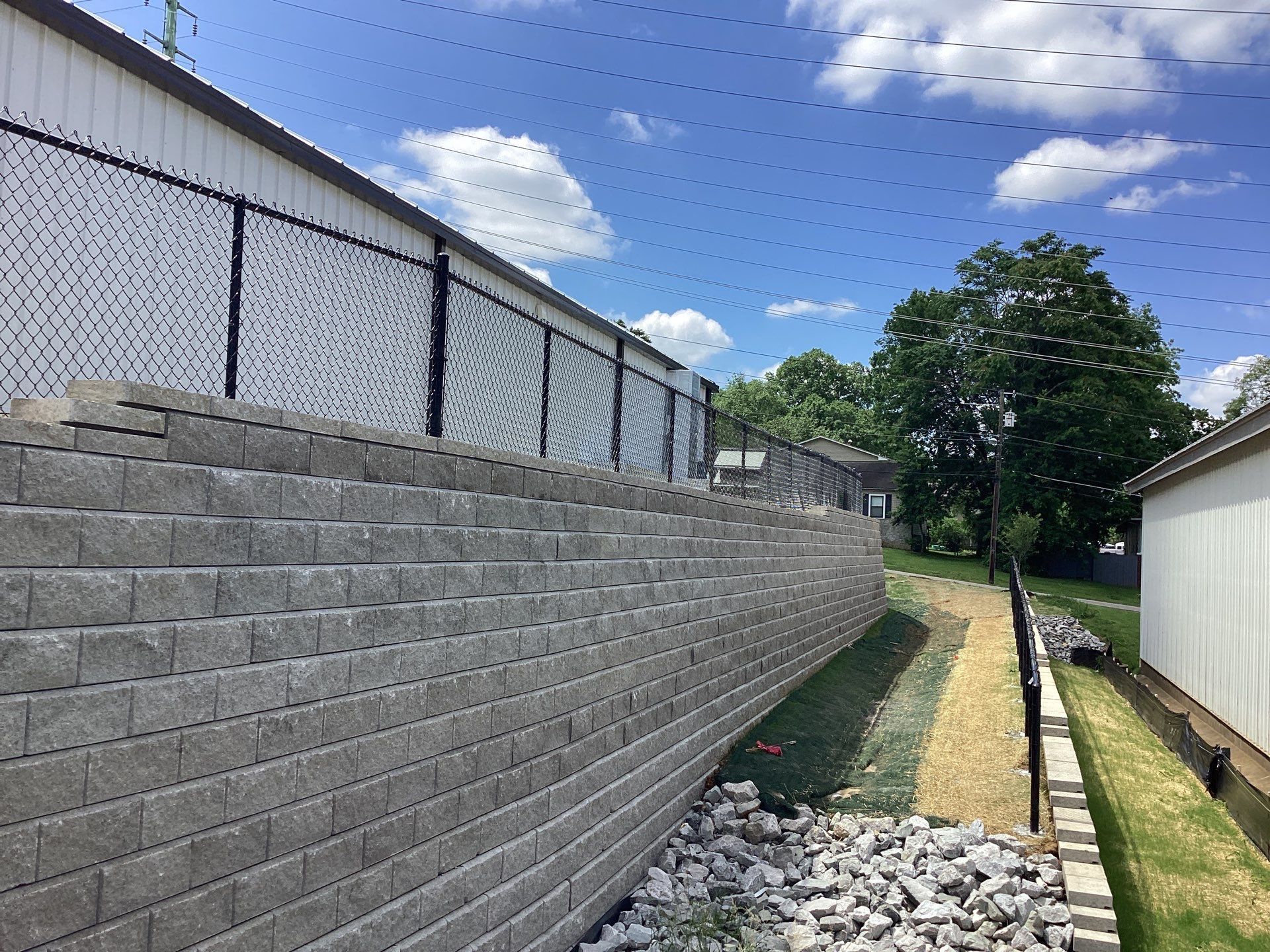 Gray retaining wall with black chain-link fence on top. Green grass, gravel, and blue sky in background.