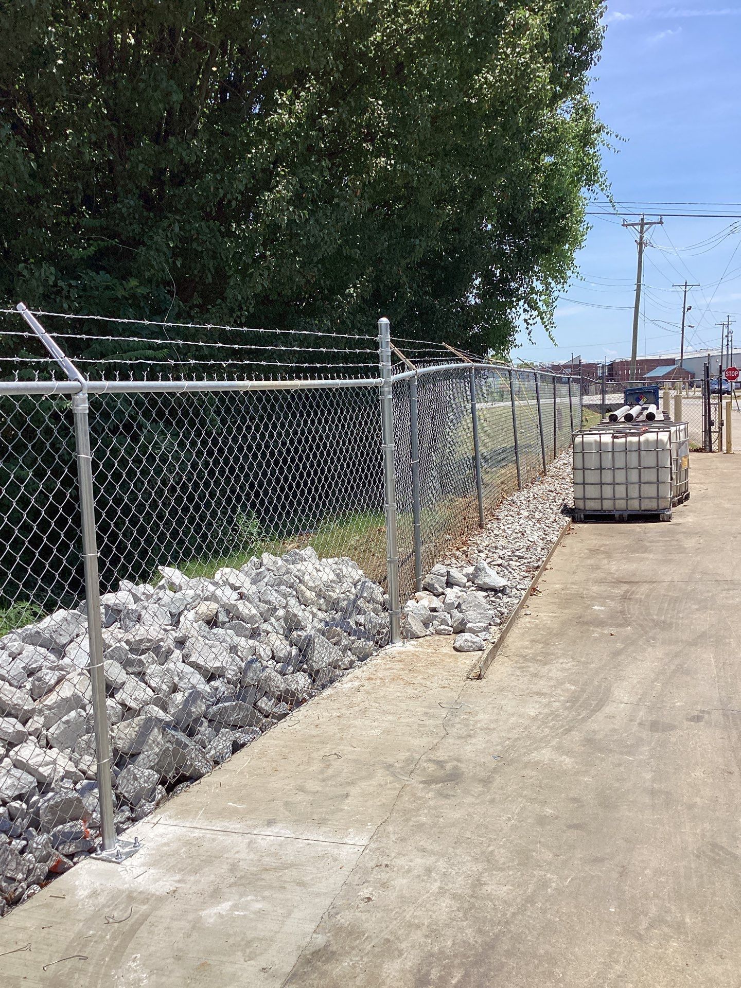 Chain link fence with razor wire, alongside concrete ground and rocks, with a tree in the background.