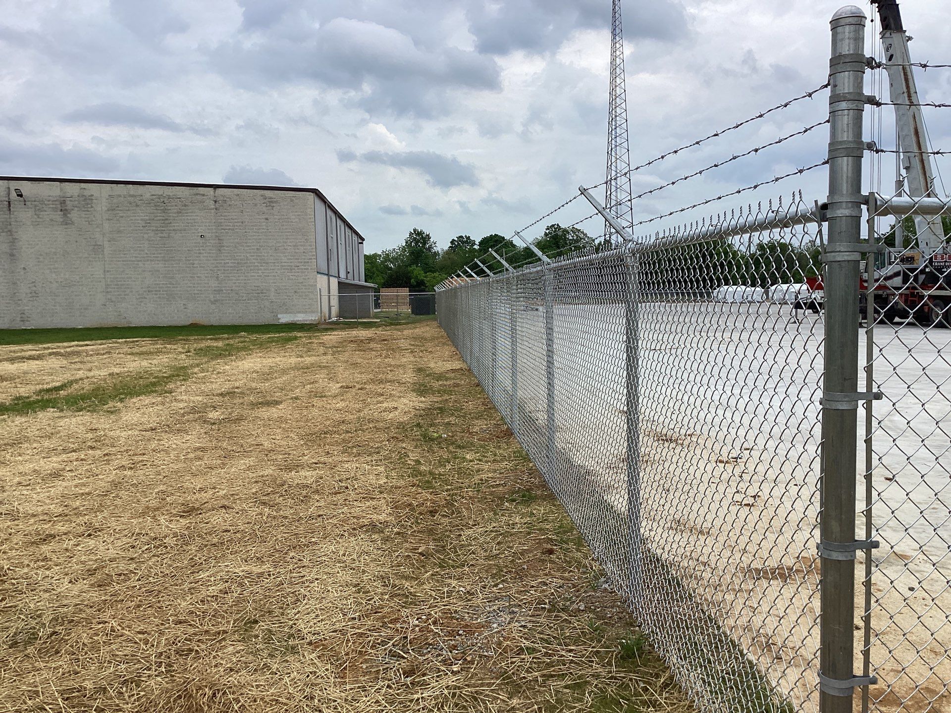 Chain-link fence with barbed wire. Field and building in background. Cloudy sky.
