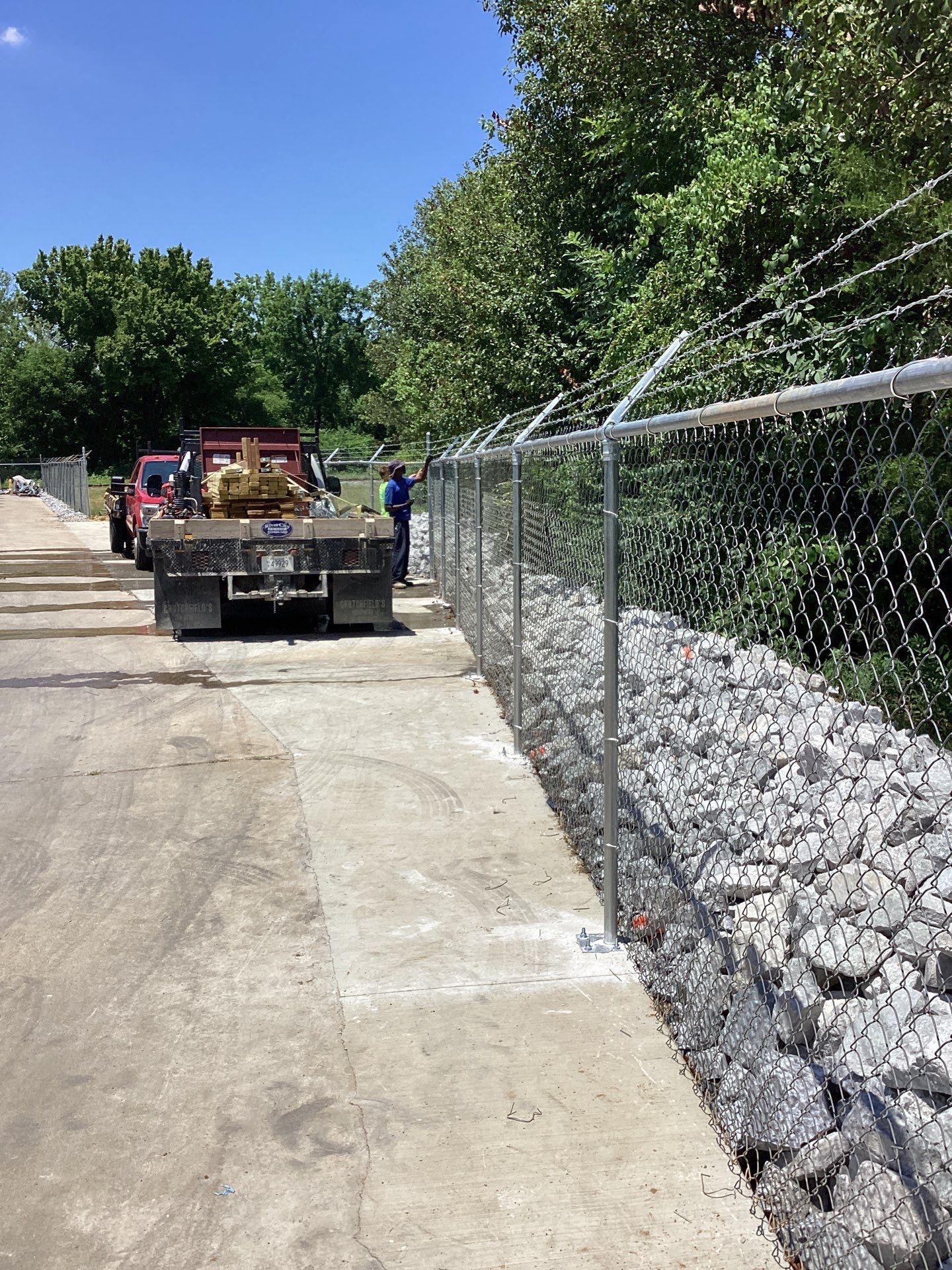 Chain-link fence with barbed wire atop a retaining wall made of stacked rocks next to a road with a truck and trees.