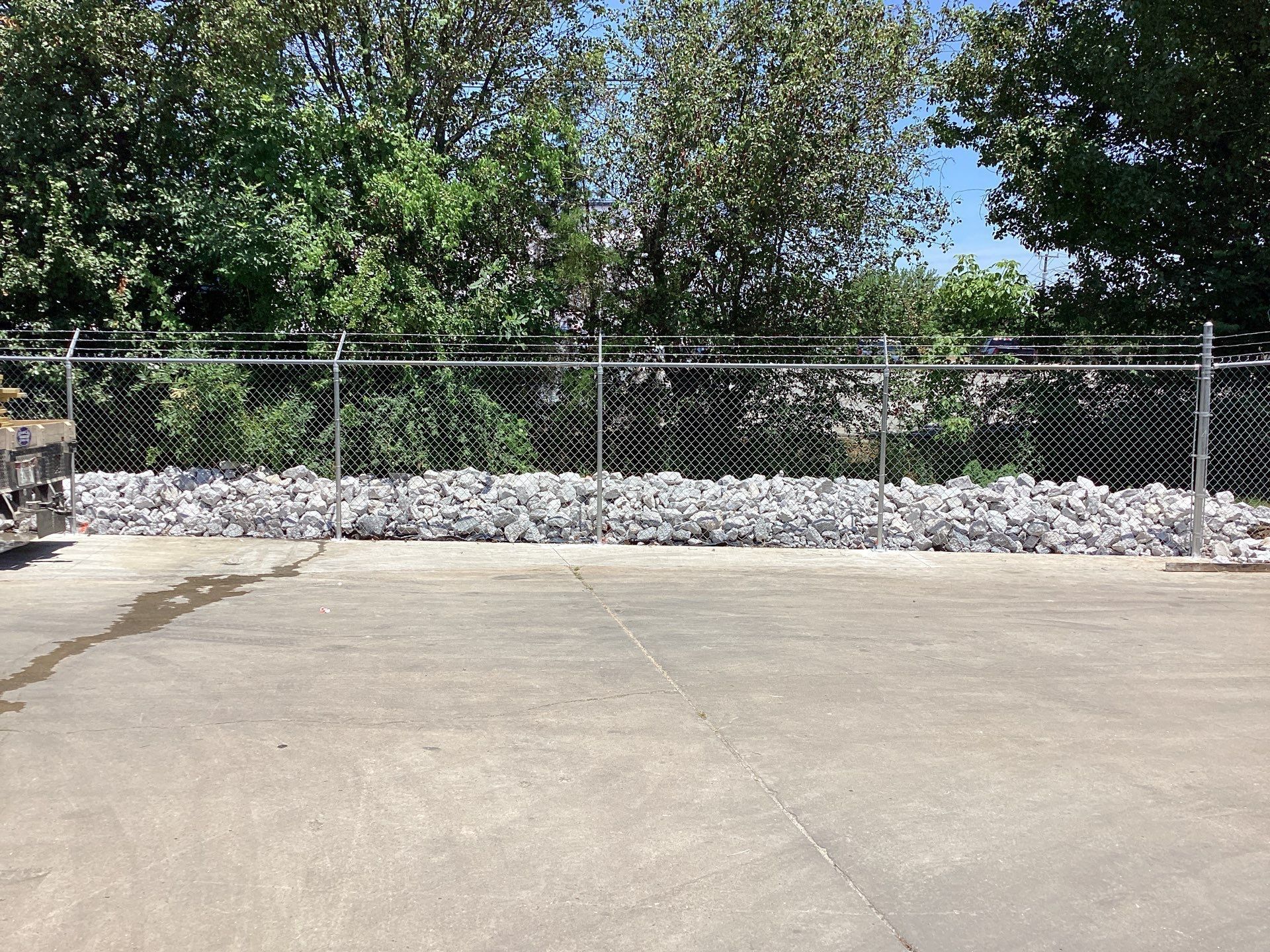 Chain-link fence with a pile of rocks at its base. Trees in the background. Paved ground in front.