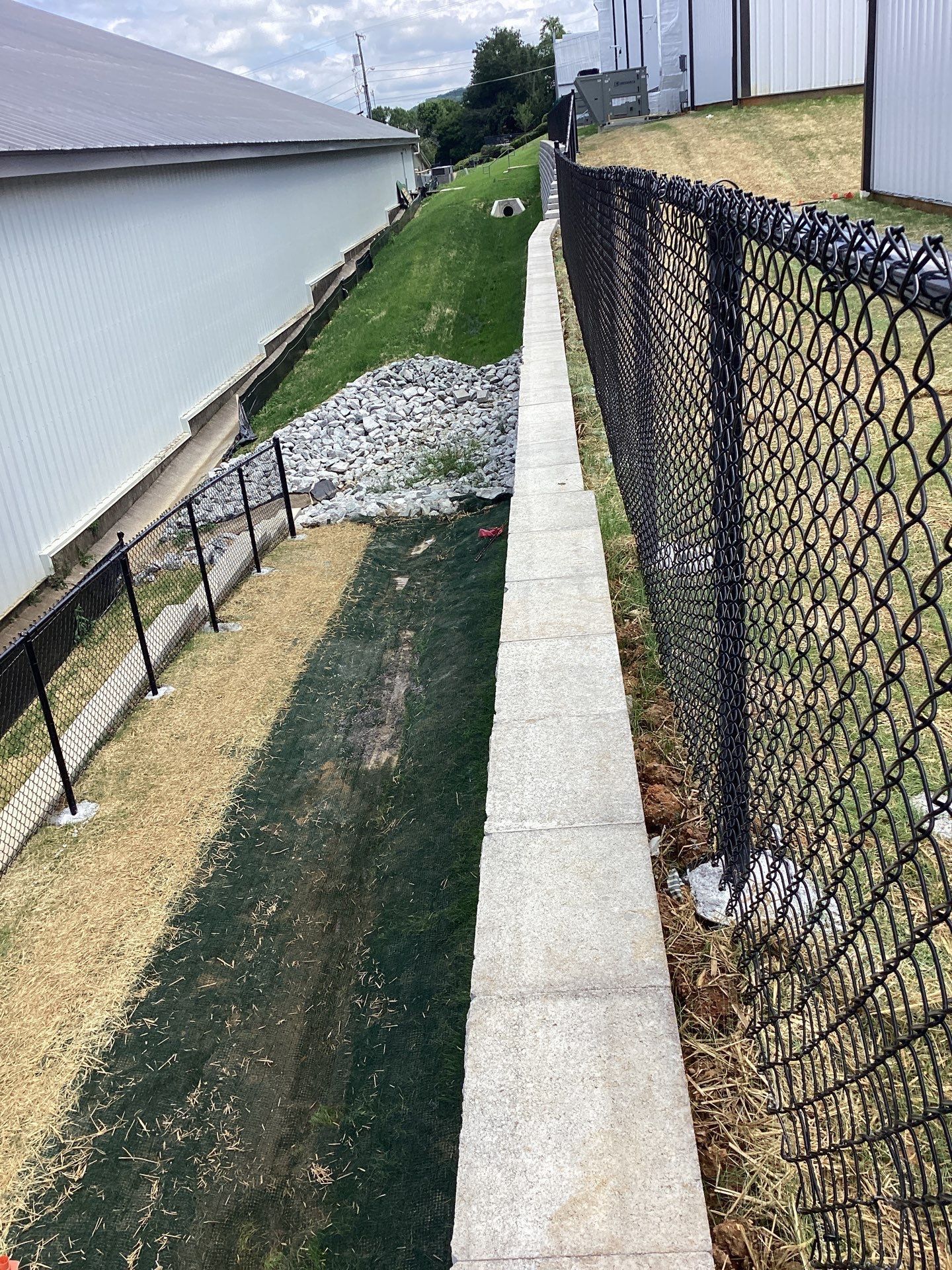 Chain-link fence borders a concrete walkway and grassy area next to a building and sloped hill.
