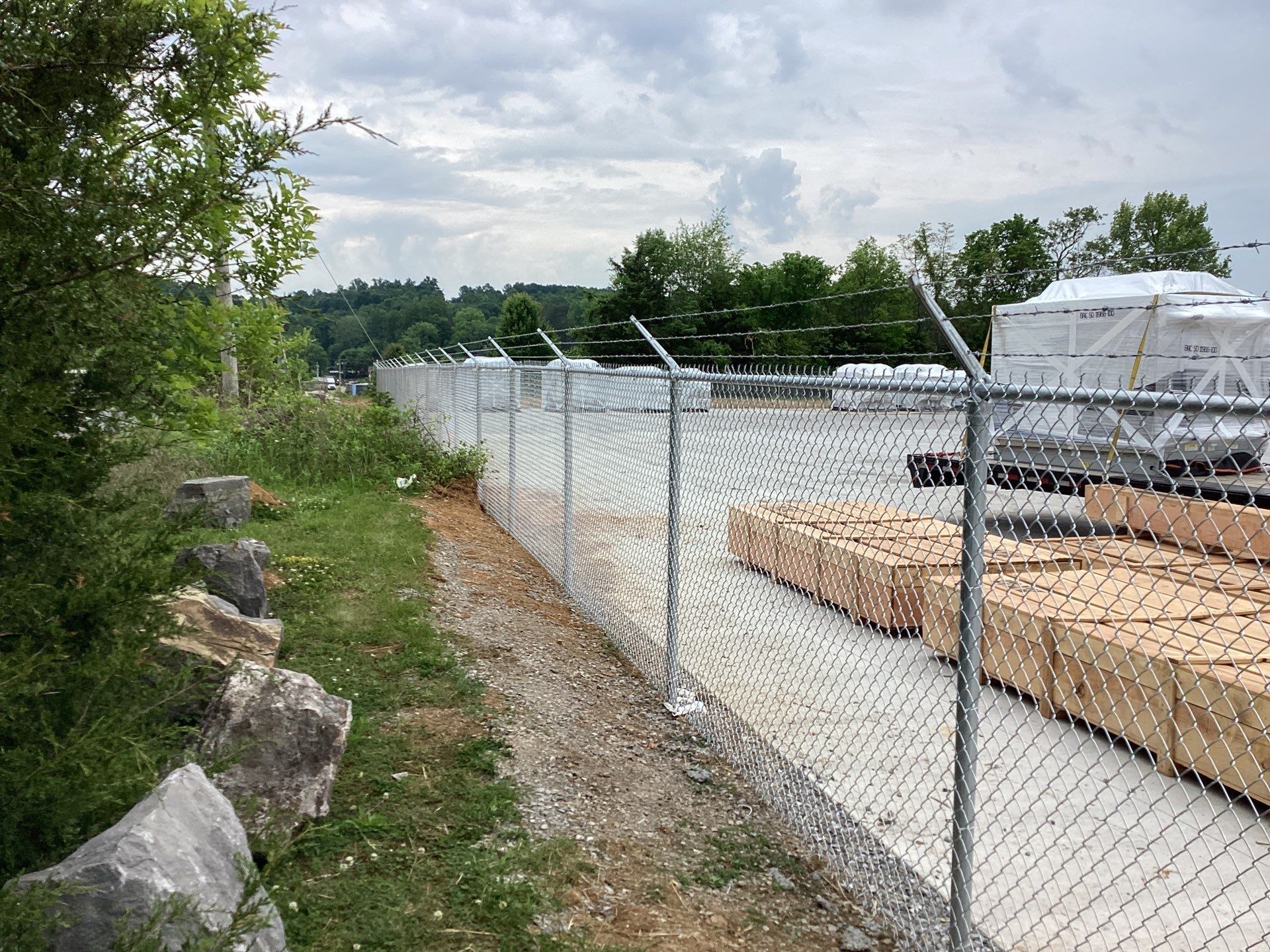 Chain-link fence with barbed wire. Timber and supplies are behind the fence. Cloudy sky, trees.