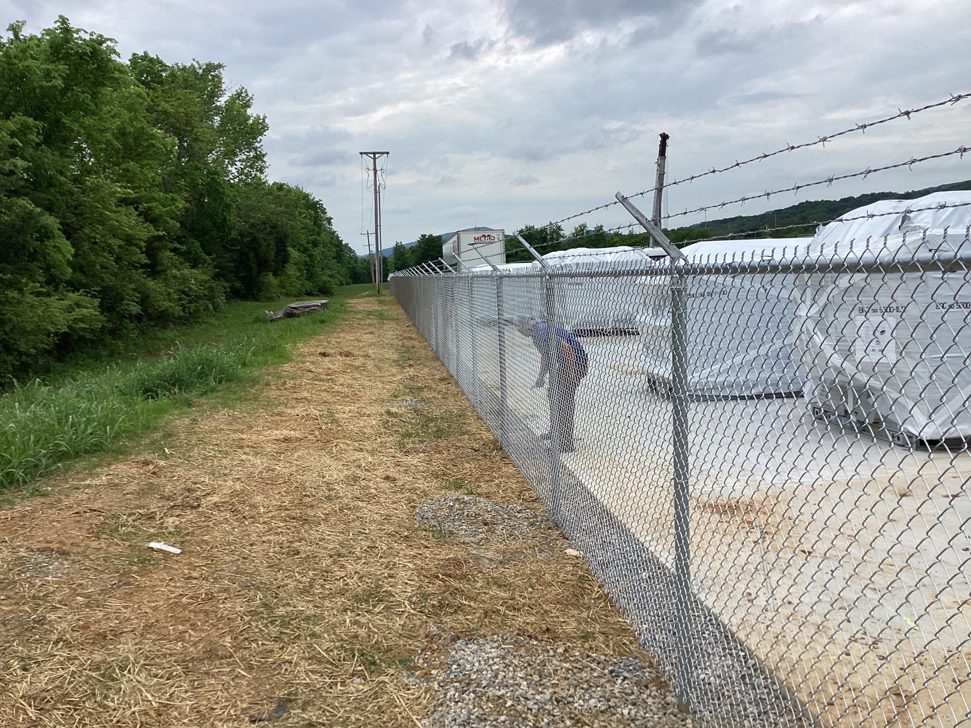 Chain-link fence topped with barbed wire runs along a grassy area, with trees on the left and storage on the right.