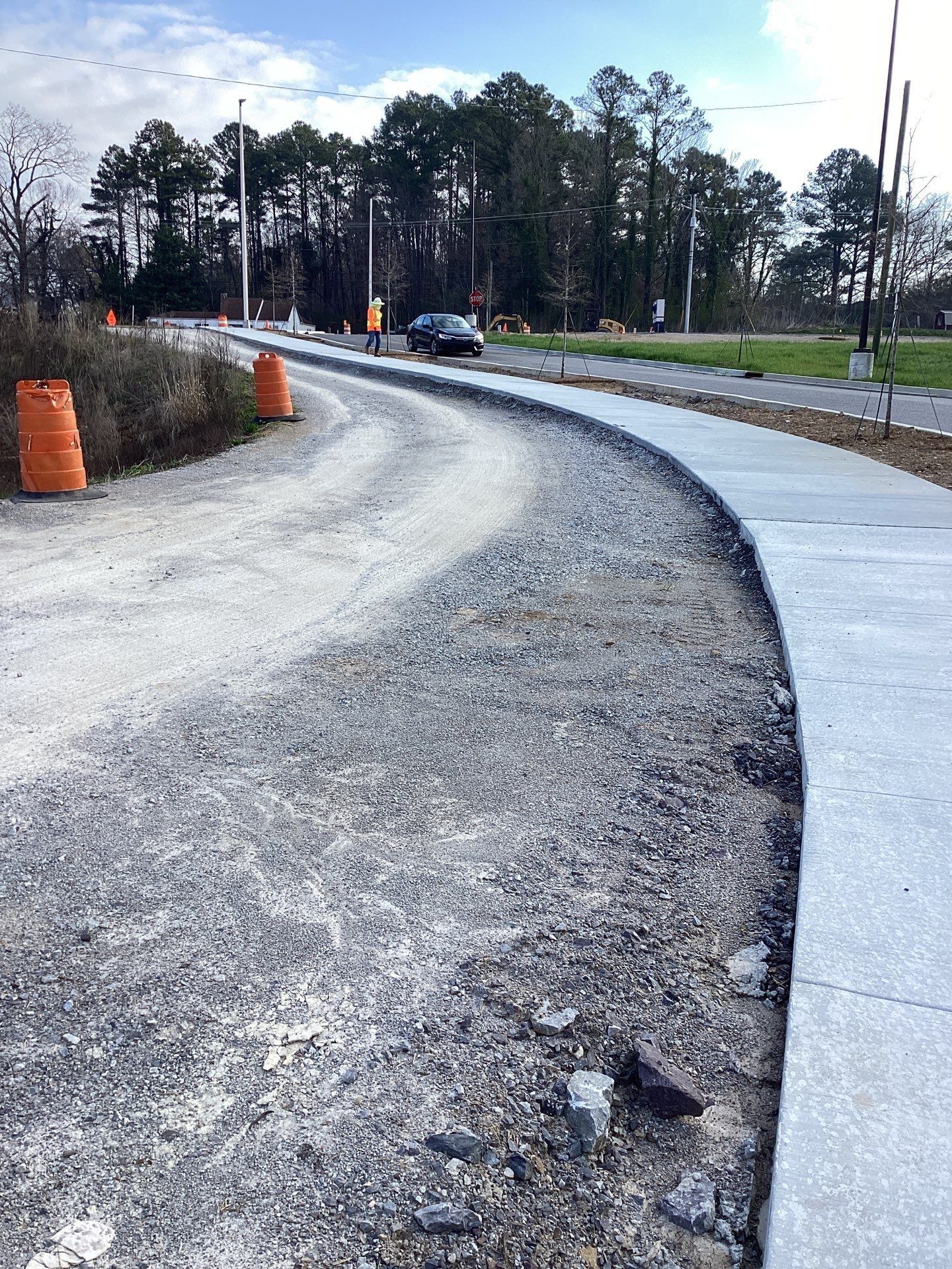 A gravel road curves alongside a new concrete sidewalk, with orange cones marking the construction site.