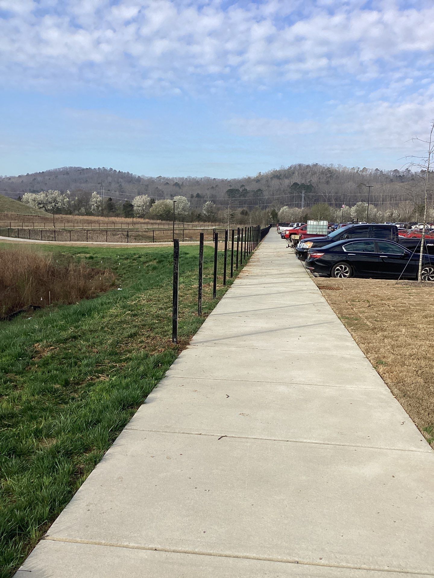 Concrete walkway bordered by grass and fence, leading towards parked cars and distant hills under a partly cloudy sky.
