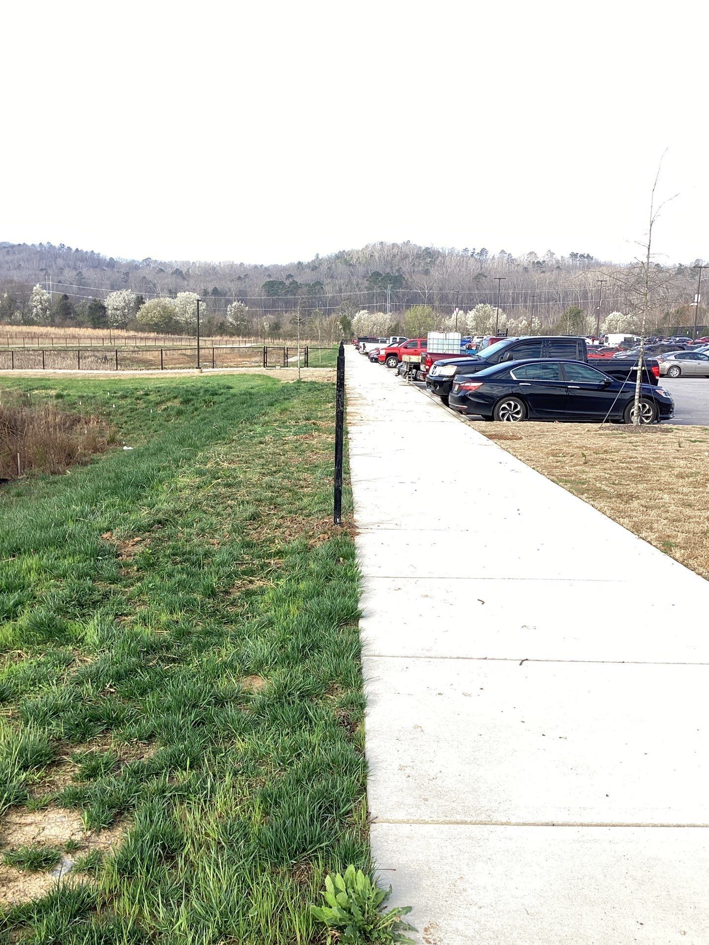 Sidewalk next to a grassy area and a parking lot with cars, mountains in the distance.