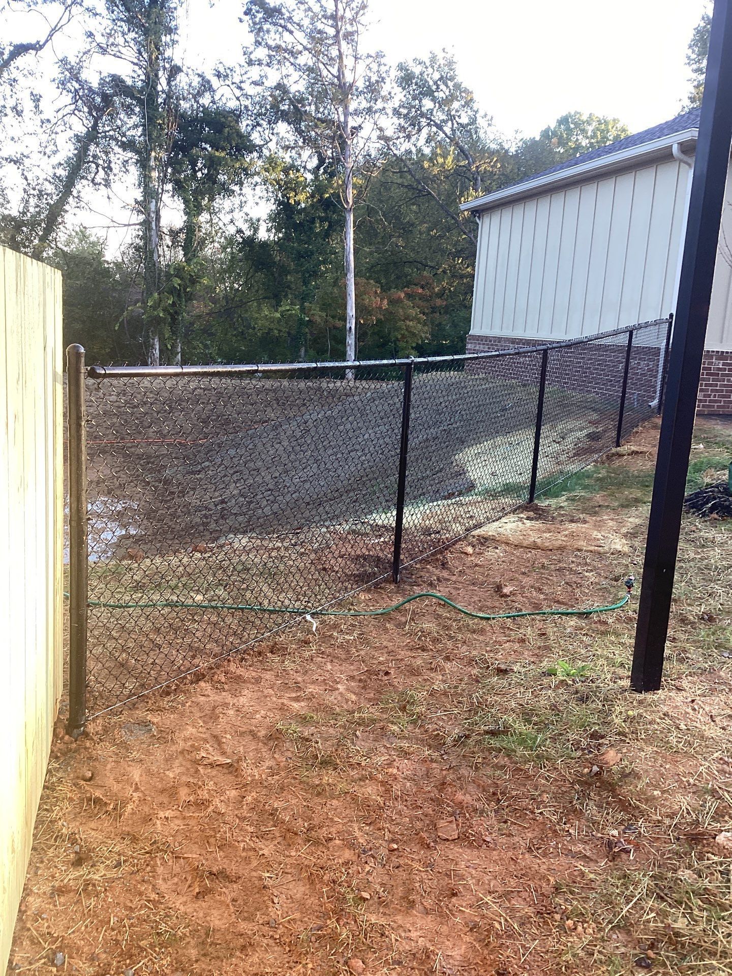 Chain-link fence next to a wooden fence. The scene is outdoors, near a building and trees, with brown dirt.