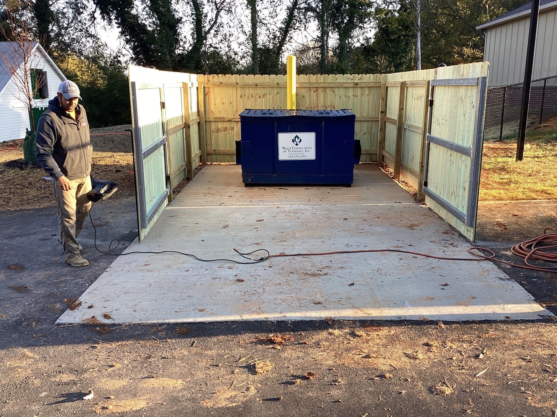 A person stands near a blue dumpster enclosed by a wooden fence, on a concrete pad.