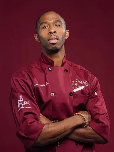 Chef in maroon coat with crossed arms, red background.