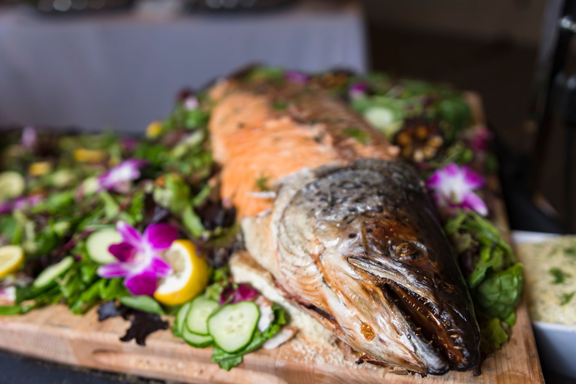 Roasted whole salmon on wooden board, surrounded by salad and lemon slices.