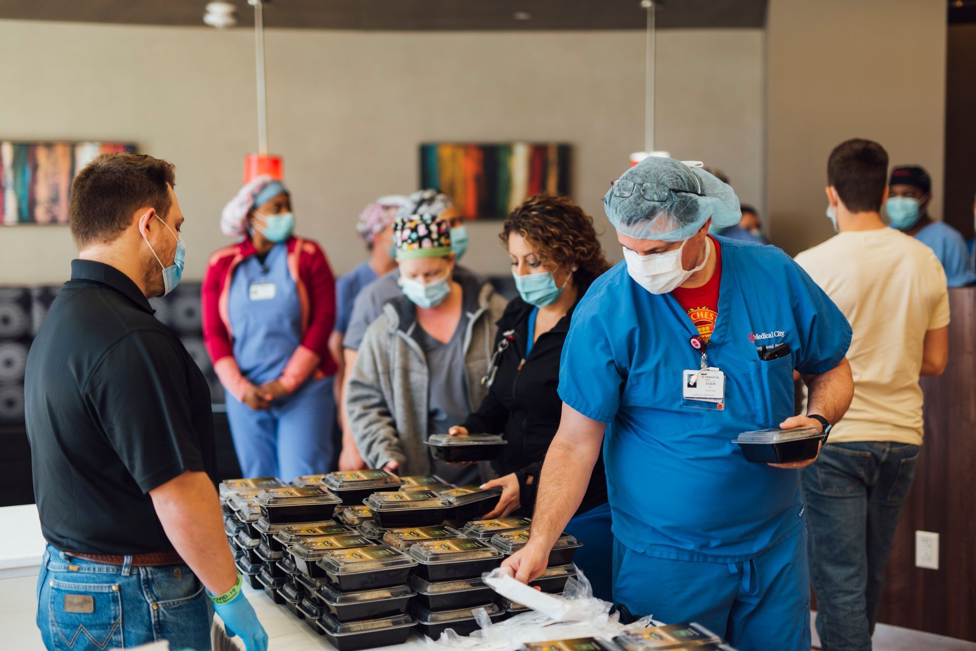 People in medical attire and masks receiving meals from a person. Other people in the background.
