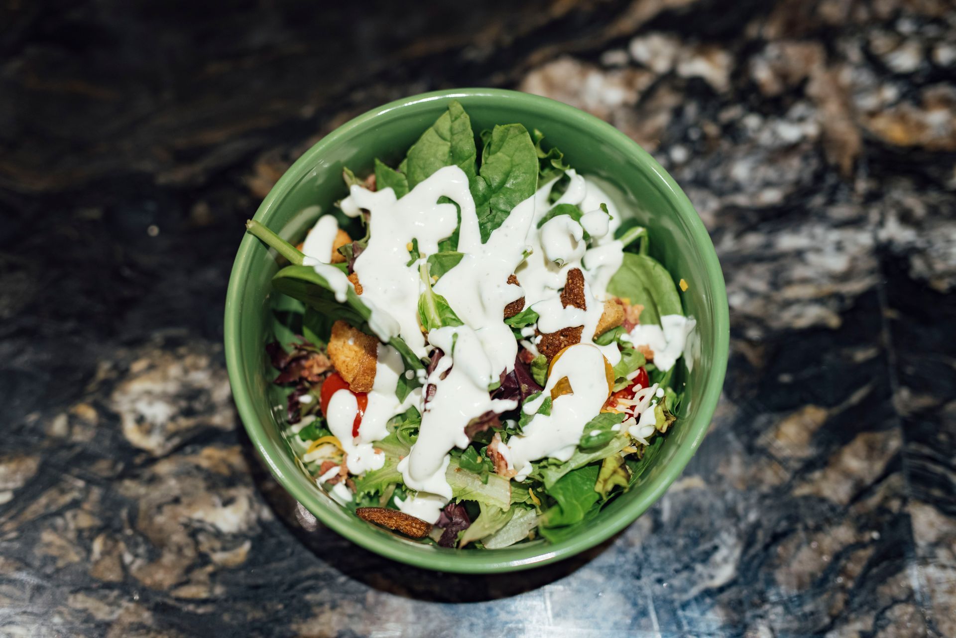 Green bowl filled with salad, topped with white dressing and bacon bits, on a marbled surface.