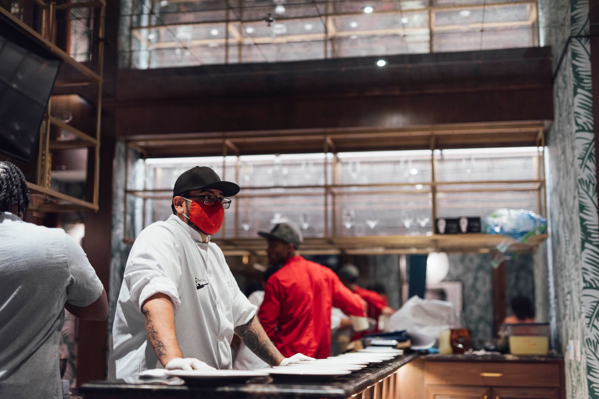Chef wearing a mask and gloves prepares food in a restaurant kitchen. Other staff nearby.