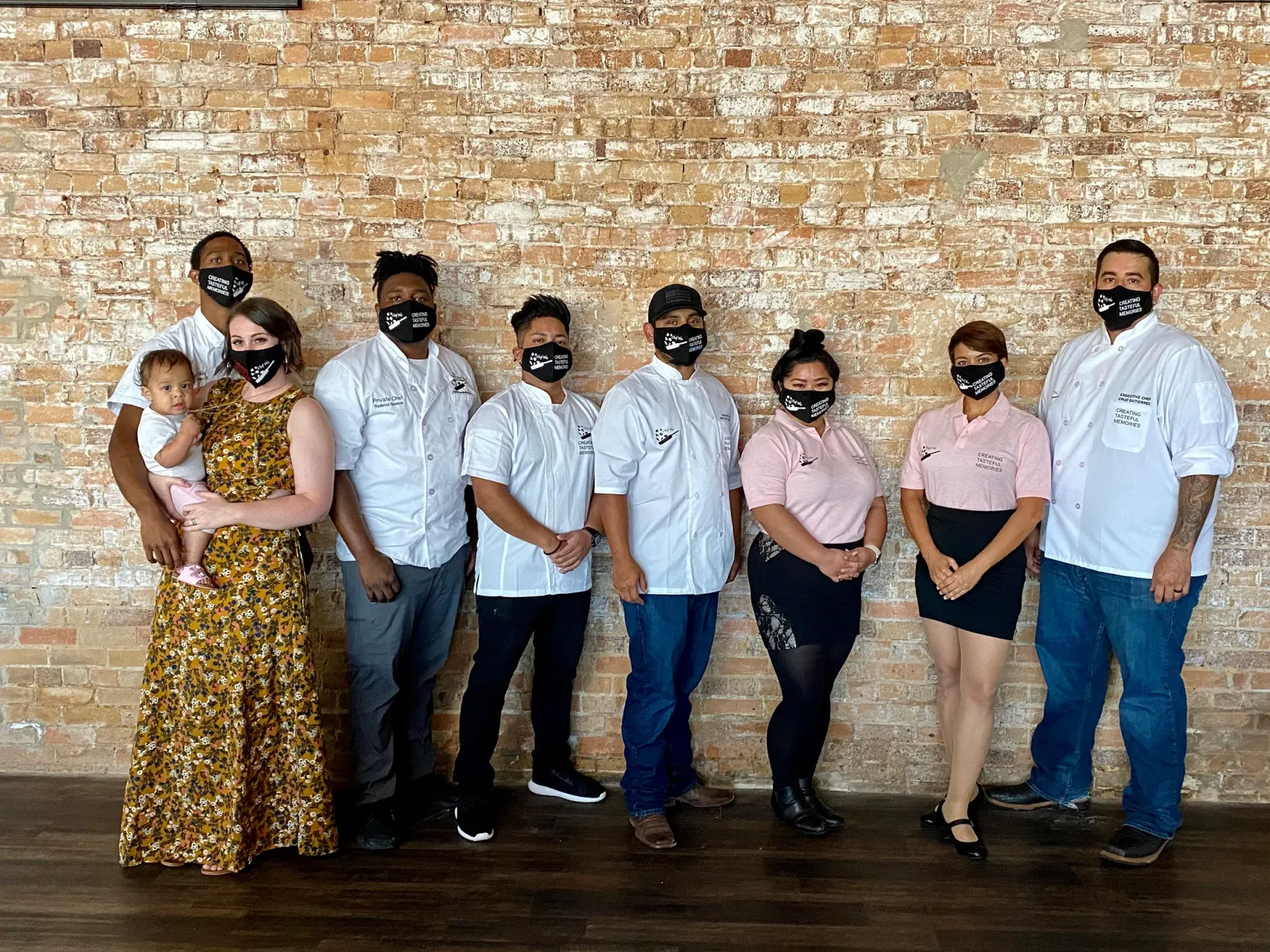 Group of restaurant staff in white shirts and masks posing in front of brick wall.