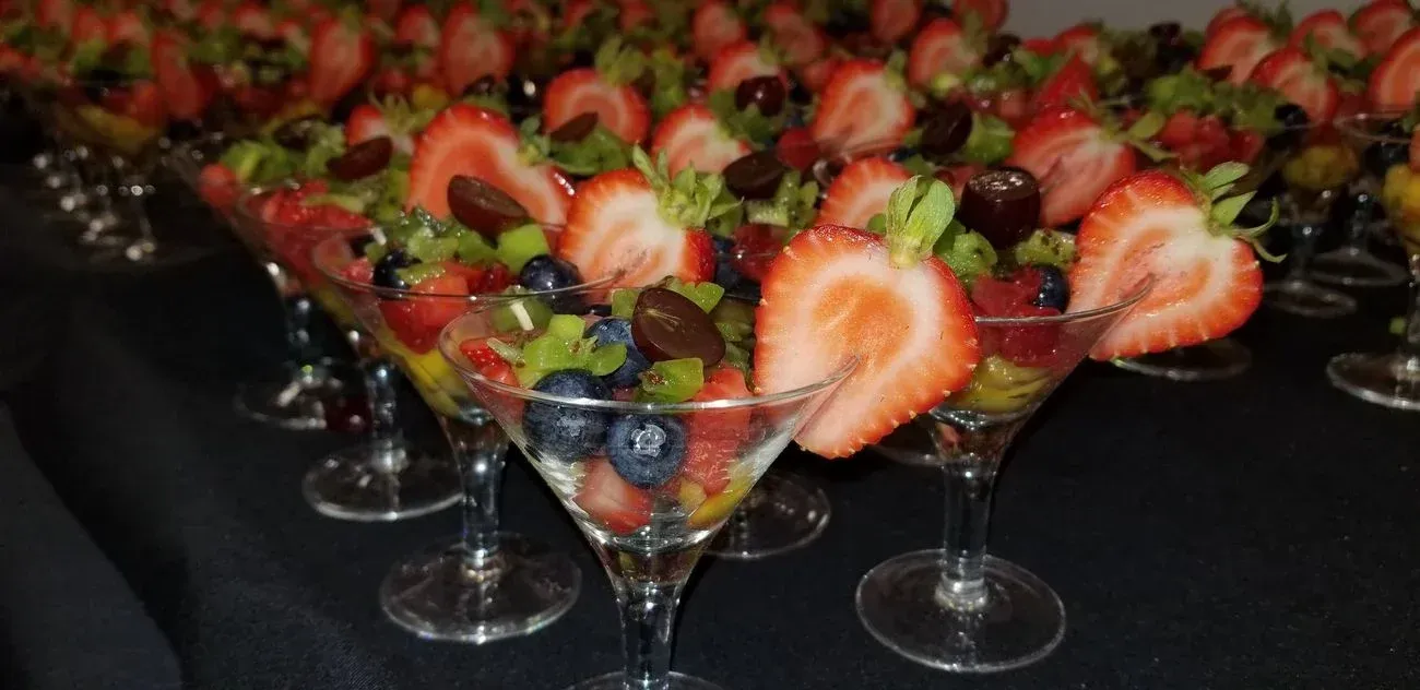 Fruit salad in martini glasses lined up on a dark surface. Strawberries, blueberries, kiwi, and grapes visible.