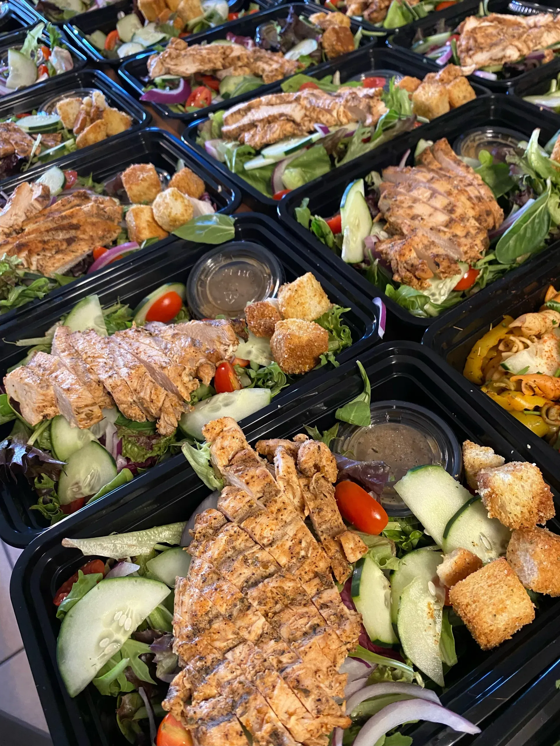 Salads in black containers with grilled chicken, cucumbers, tomatoes, and dressing, likely for takeout.