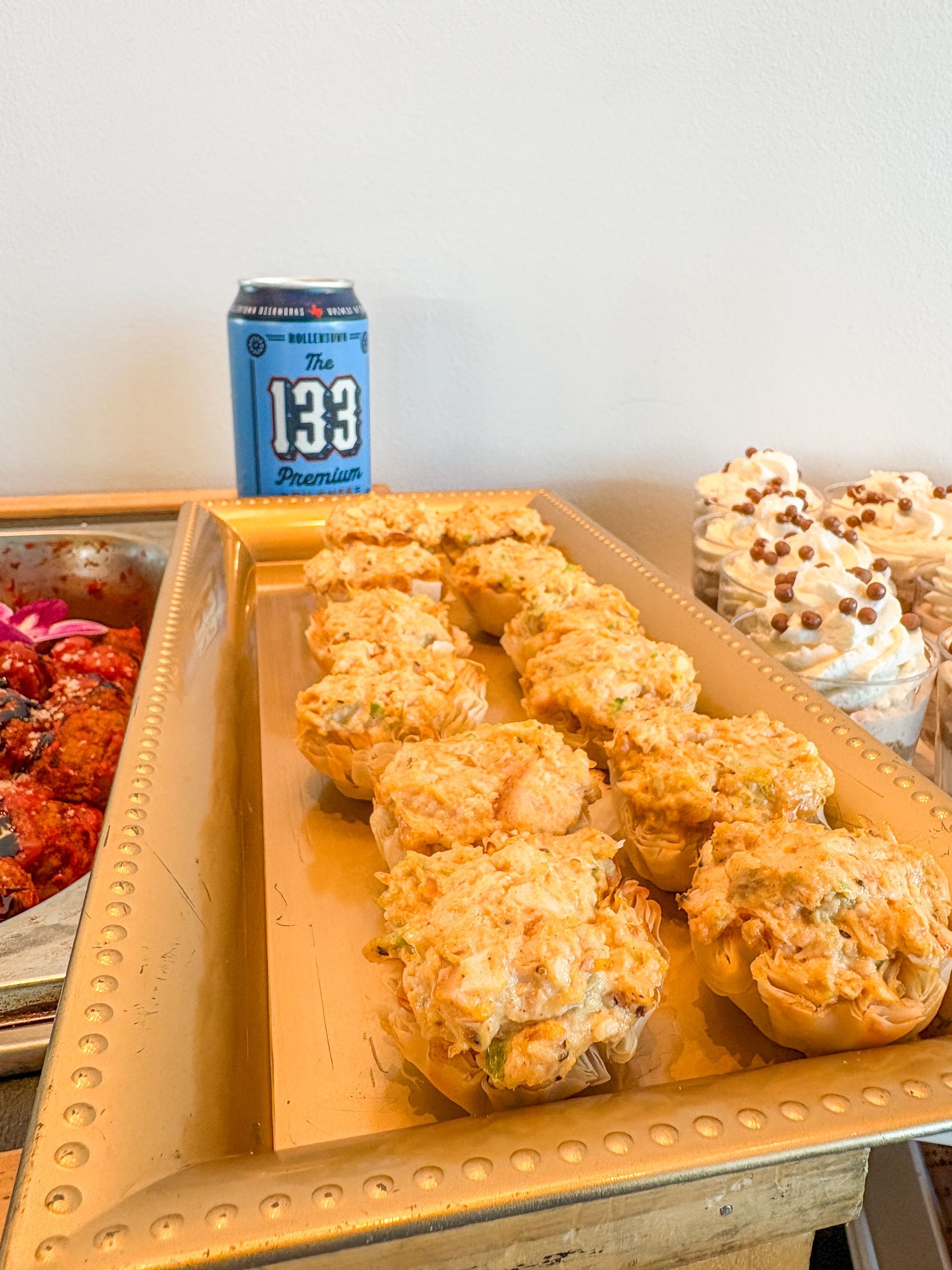 A golden tray of savory appetizers. A can of blue beer and other foods are visible in the background.