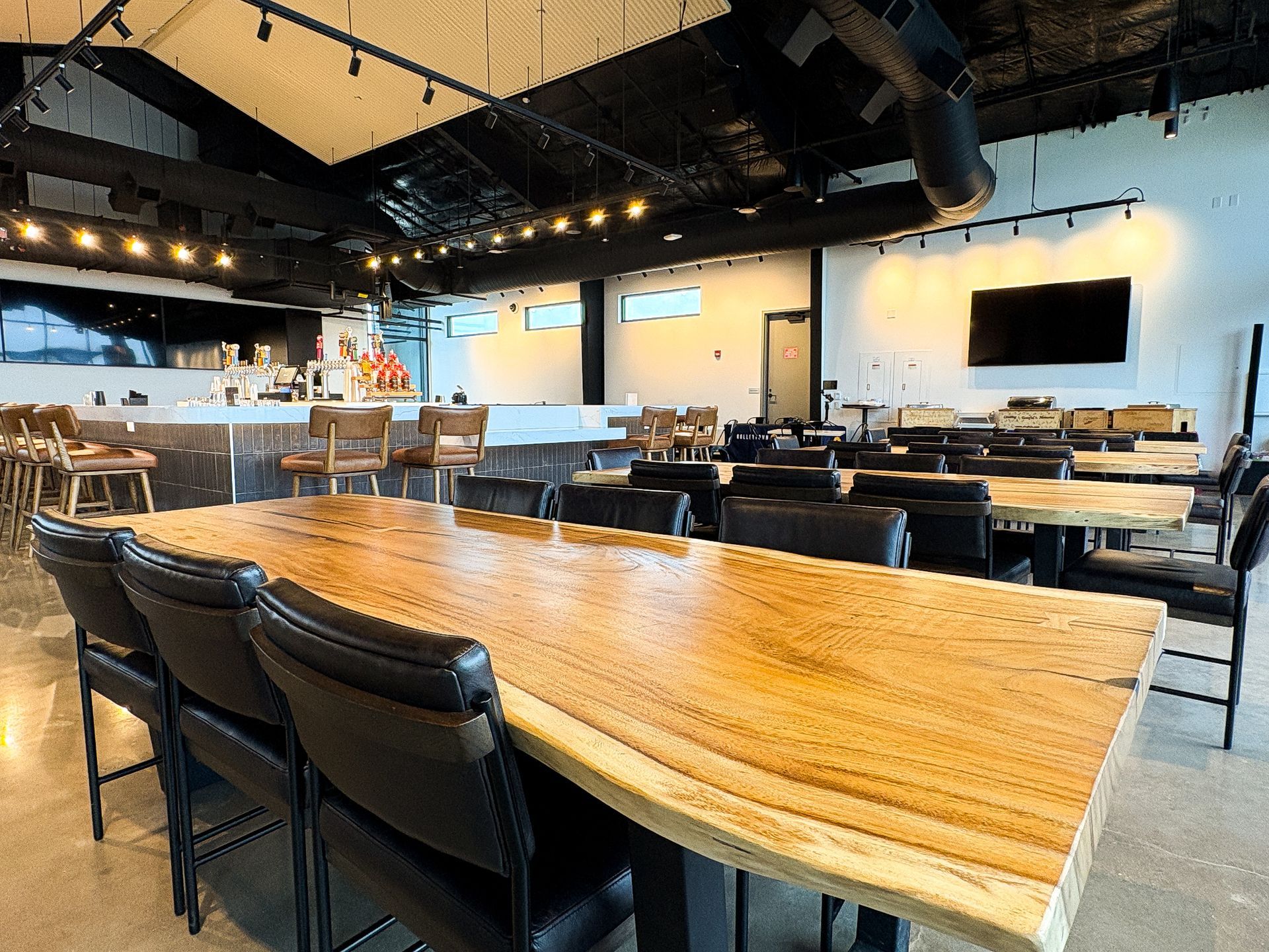Interior of a restaurant with long wooden tables, bar, and seating. Black ceiling and white walls.