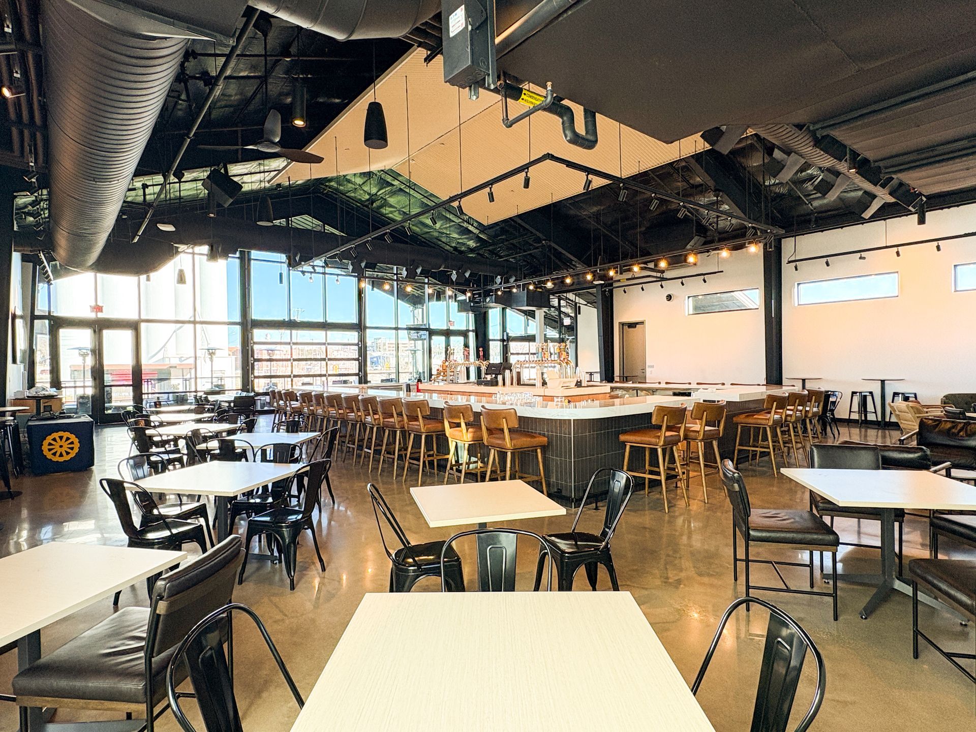 Interior of a modern bar with a long counter, tables, and chairs. Large windows and a black ceiling.