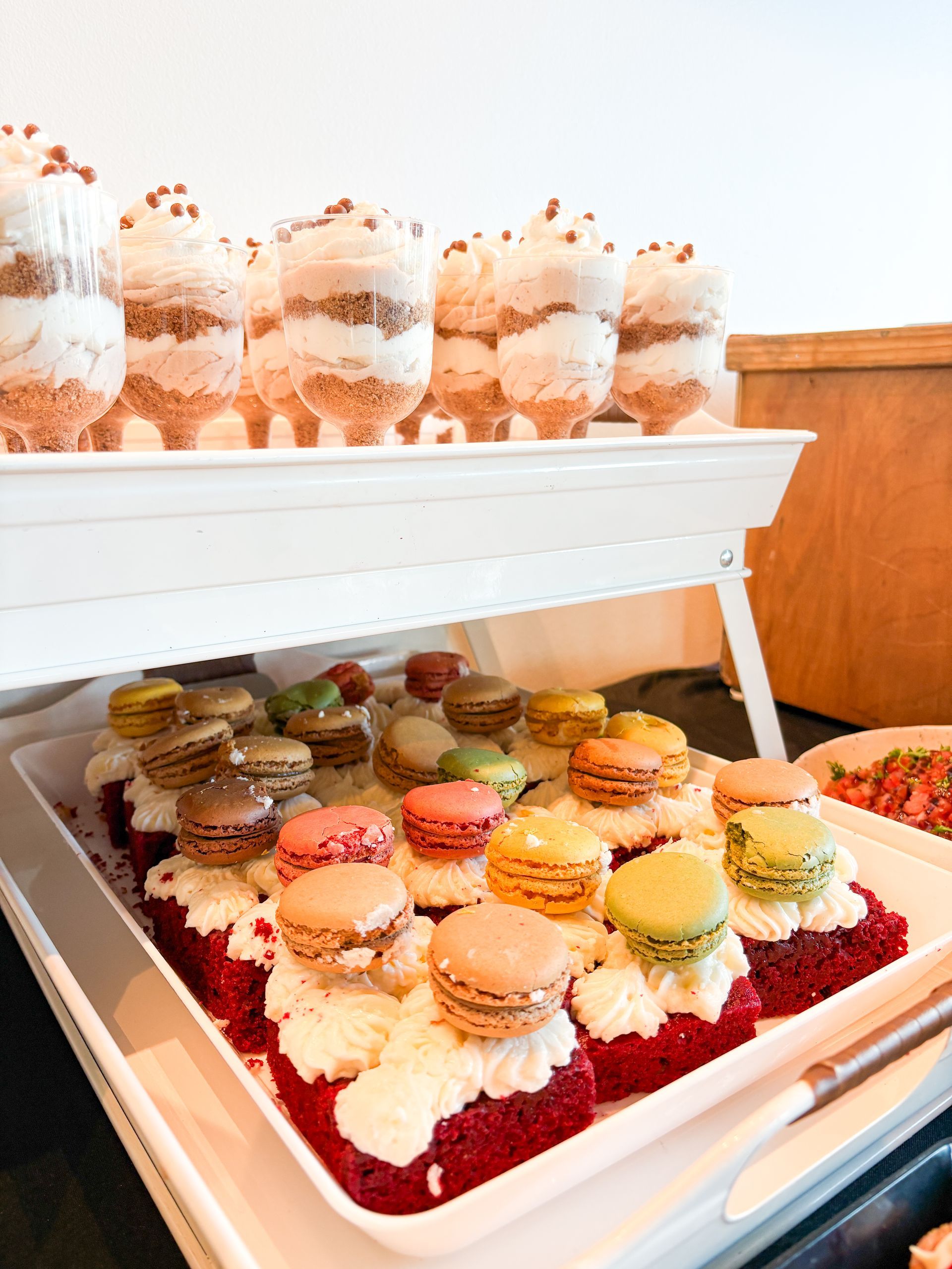 Dessert display with layered trifles and colorful macarons on red cake, on a white tiered tray.