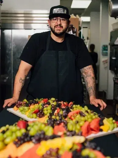 Chef in black apron and cap stands behind a table laden with colorful fruit platters.