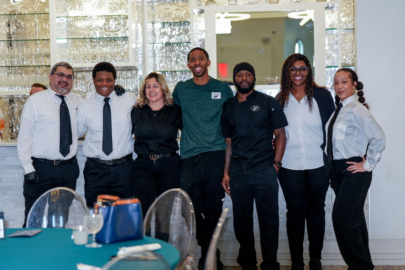 Group of restaurant staff posing for a photo, smiles, formal attire, bar in background.