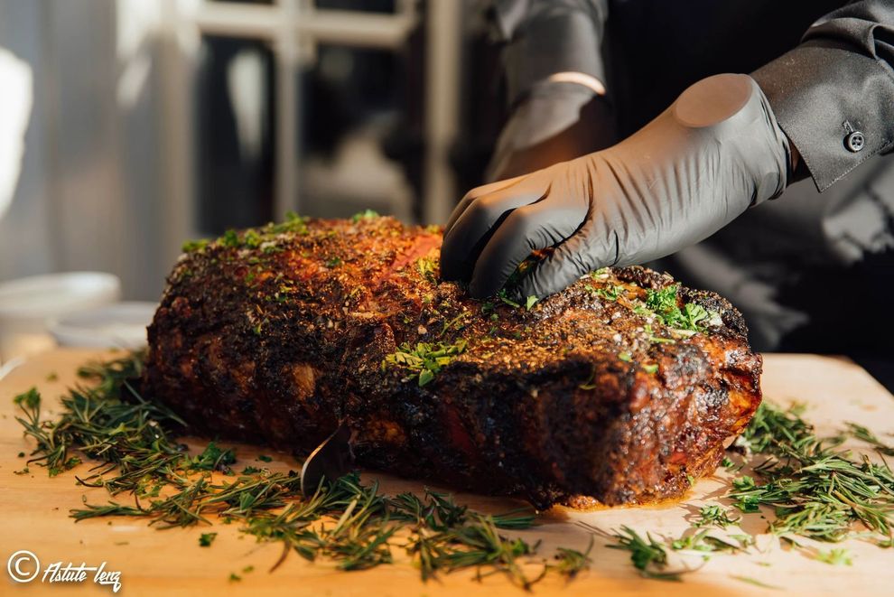 A person in black clothing and gloves prepares a large, herb-crusted roasted meat on a wooden cutting board.