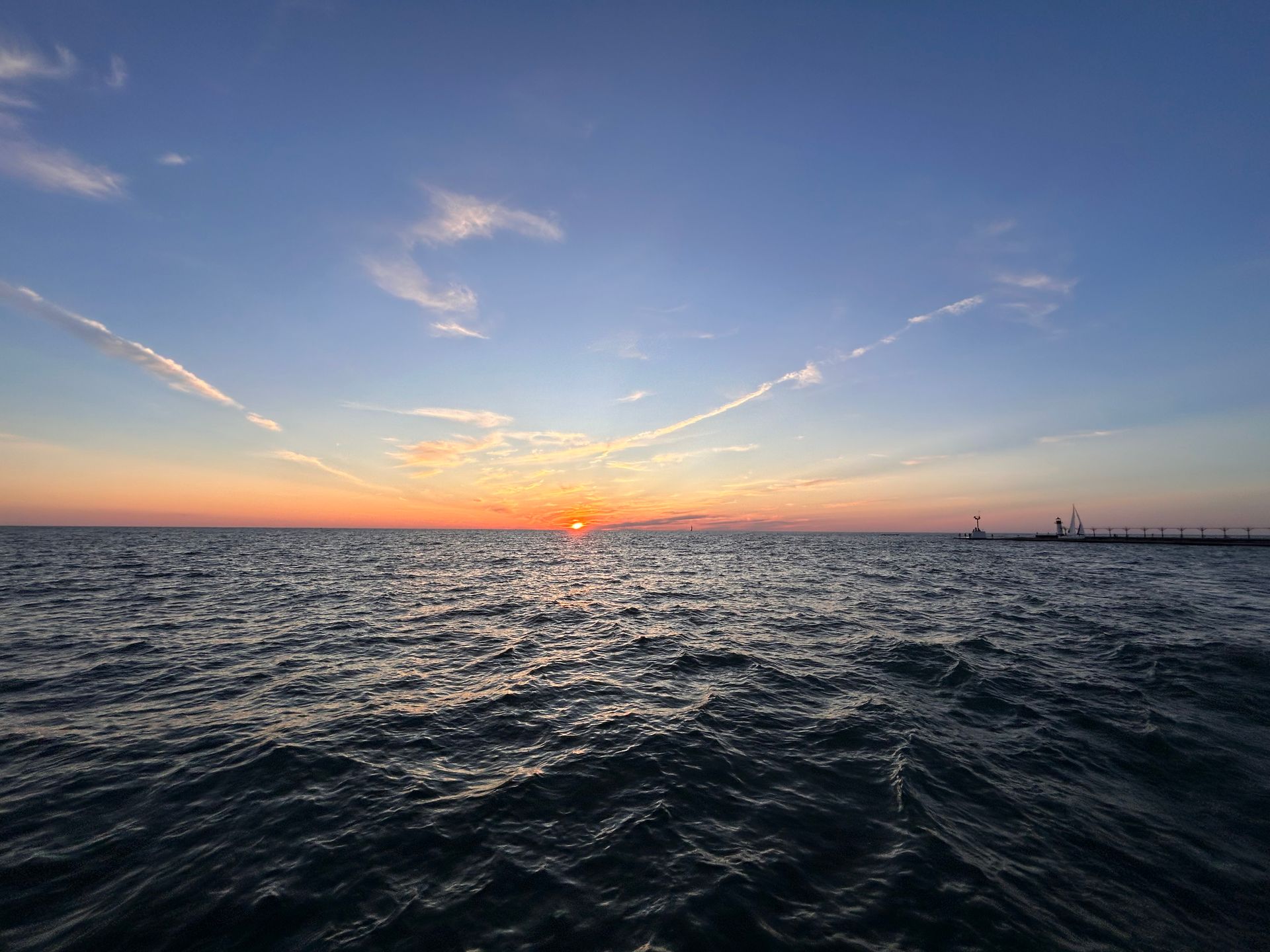 Ocean sunset with orange sun over the horizon, dark water reflecting light, blue sky with wispy clouds.